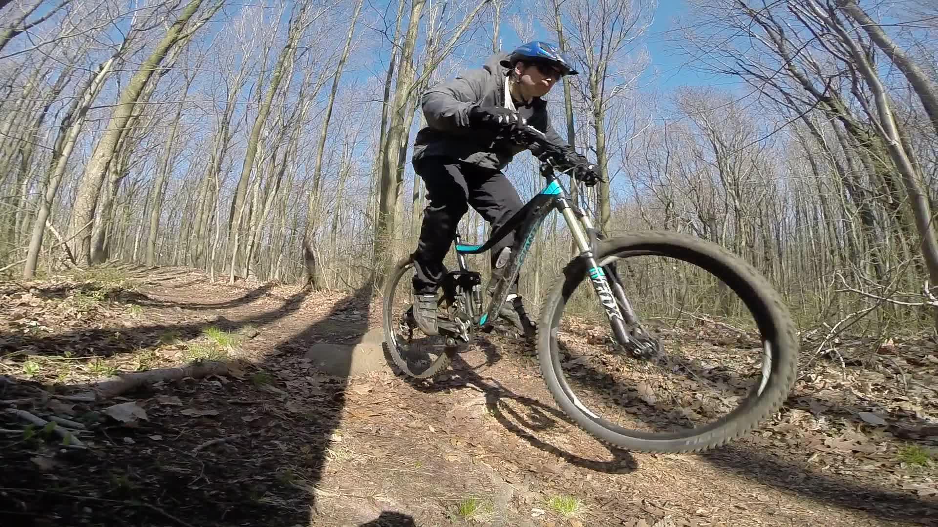 A person riding a mountain bike on a dirt trail in a forested area, with trees in the background and blue sky above. The rider is wearing a helmet and riding gear, actively navigating the trail. Richmond Avenue and Forest Hill road mountain bike trail.