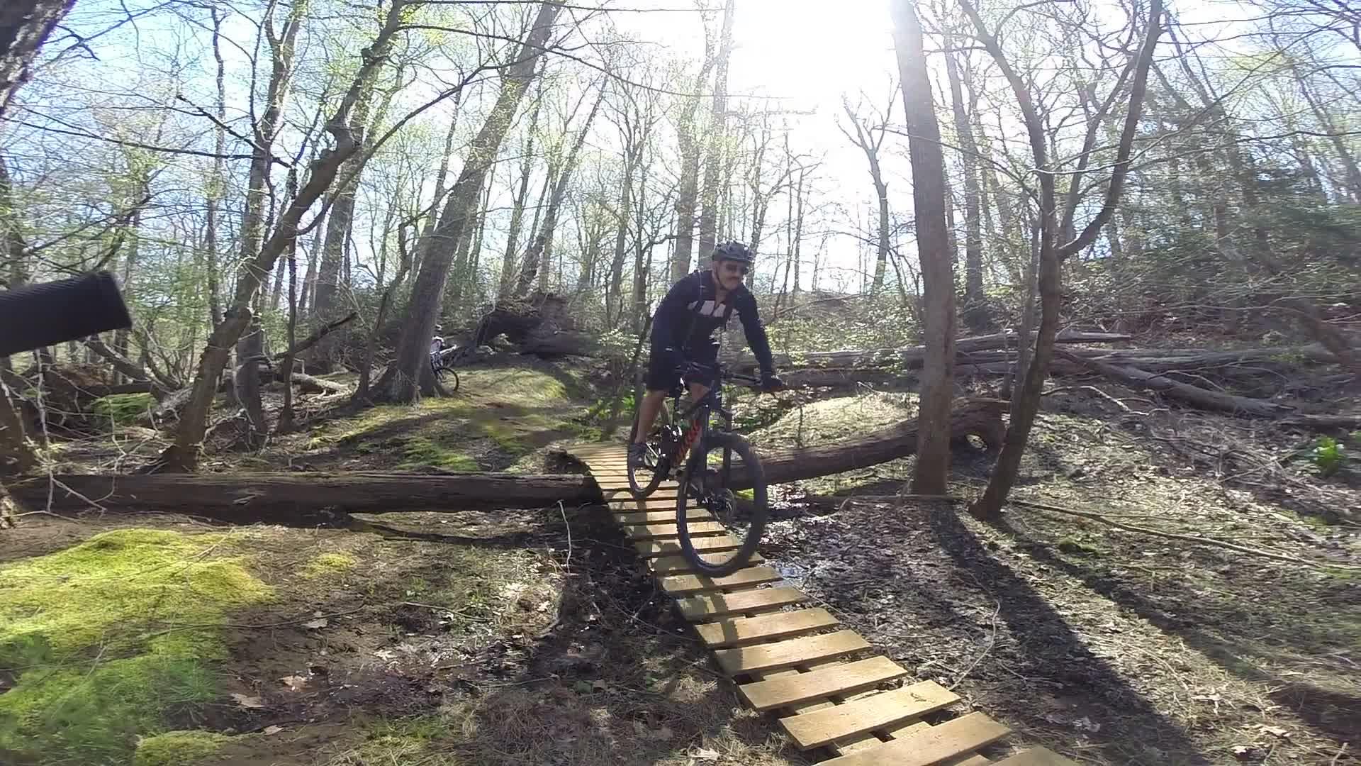 A mountain biker navigates a wooden bridge in a wooded area, surrounded by trees and greenery, with sunlight streaming through the branches. The rider is wearing a helmet and biking gear, showcasing a moment of skillful balance on the narrow path. Allaire State Park mountain bike trail.