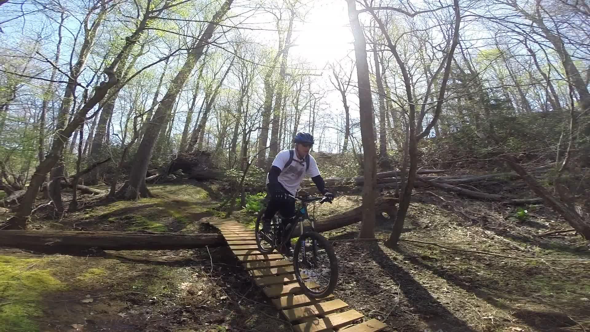 A person riding a mountain bike on a wooden bridge in a wooded area. Sunlight filters through the trees, highlighting the greenery and the natural landscape. Allaire State Park mountain bike trail.