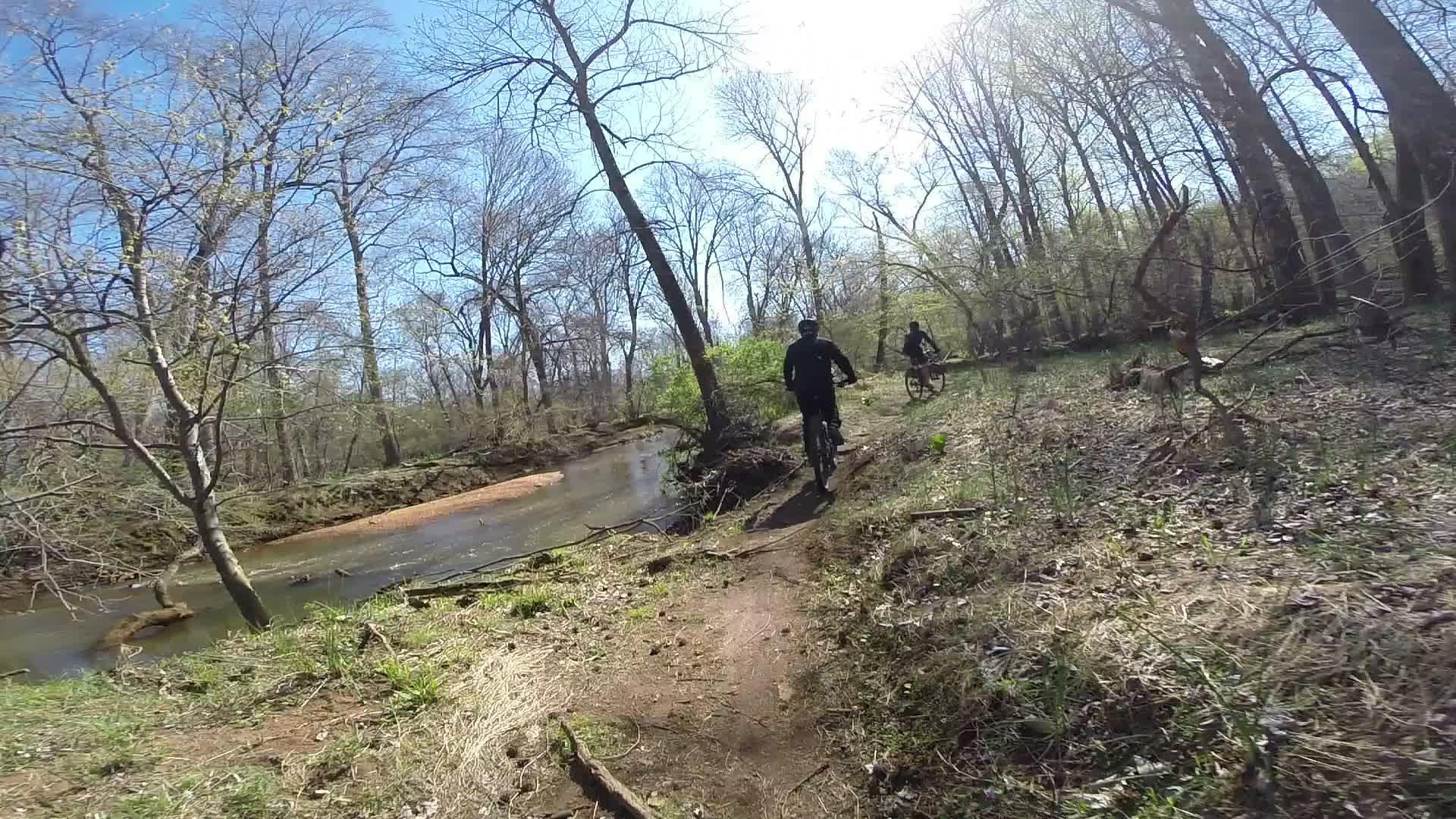 Two mountain bikers ride along a narrow dirt path beside a creek, surrounded by trees and greenery under a clear blue sky. Sunlight filters through the branches, creating a bright and inviting outdoor scene. Allaire State Park mountain bike trail.