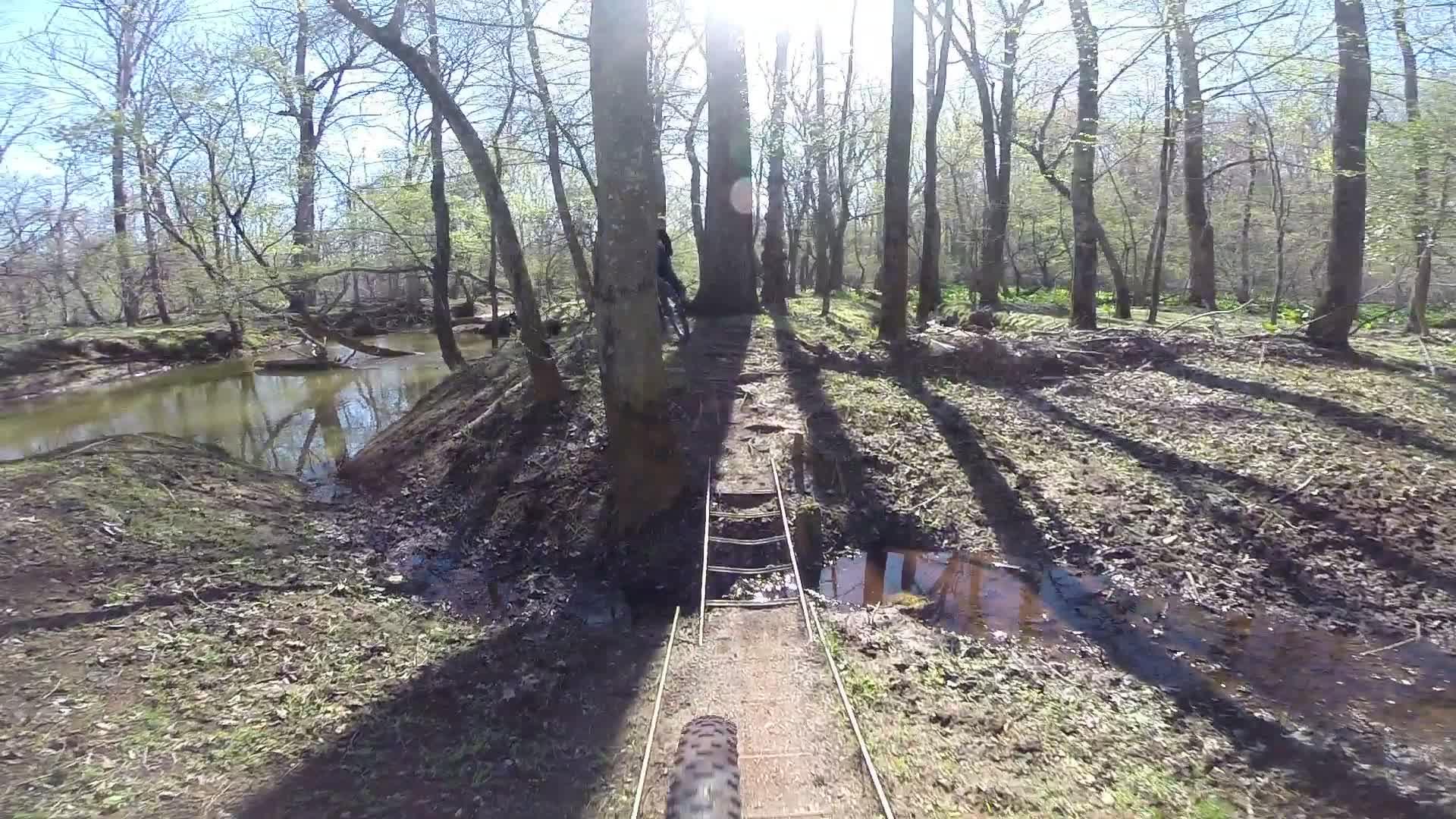 A narrow footpath winding through a sunlit forest, bordered by tall trees and shadows. A small creek flows nearby, with a rustic wooden bridge crossing a muddy area. Bright green foliage is visible in the background, indicating springtime. Allaire State Park mountain bike trail.
