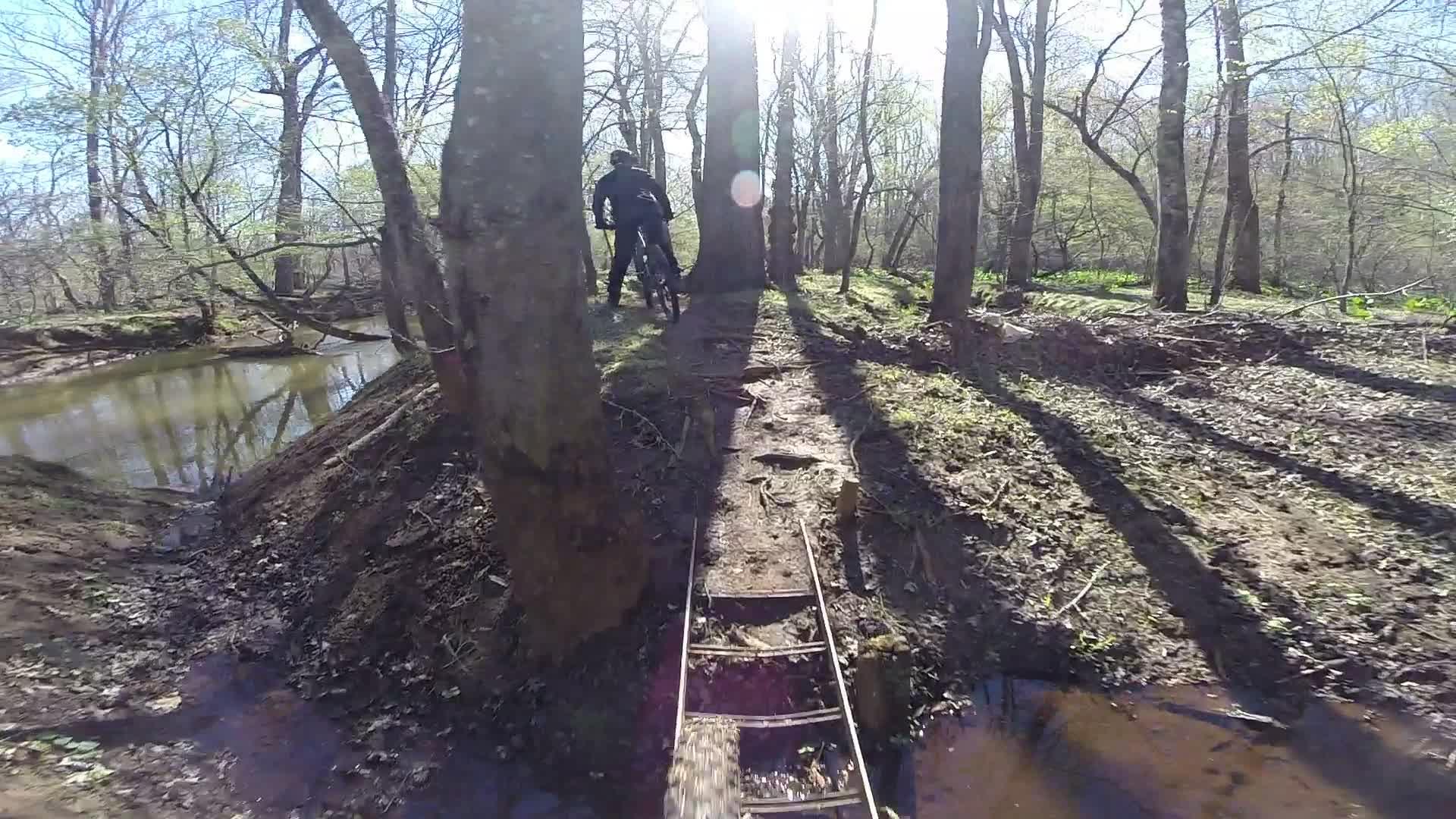 A mountain biker navigating a narrow dirt path through a wooded area, with trees casting long shadows on the ground and sunlight filtering through the branches. A calm stream is visible nearby, reflecting the surrounding nature. Allaire State Park mountain bike trail.