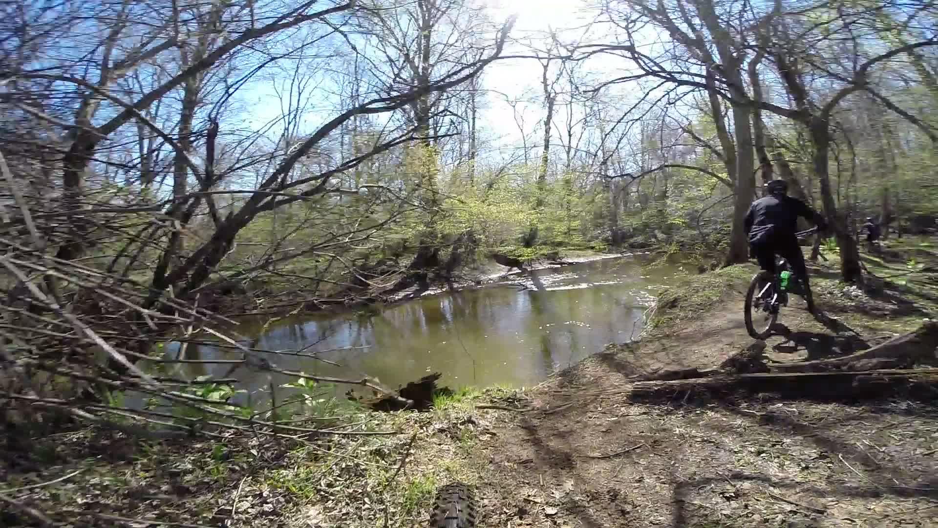 Mountain biker riding along a dirt trail beside a serene creek, surrounded by leafless trees and budding greenery. The sun shines brightly in the blue sky, casting reflections on the water. Allaire State Park mountain bike trail.
