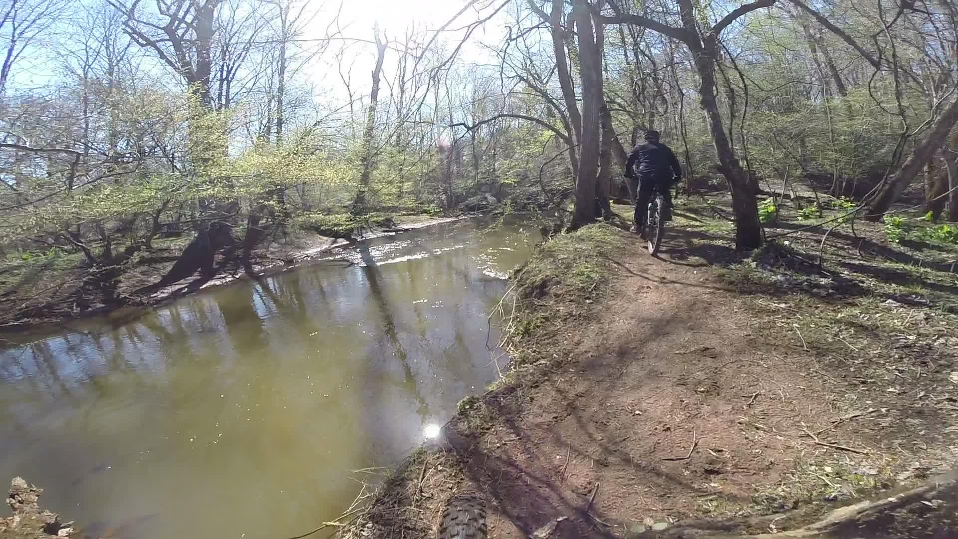 A cyclist riding along a narrow path next to a calm body of water, surrounded by trees in early spring. Sunlight filters through the branches, casting reflections on the water's surface. Allaire State Park mountain bike trail.