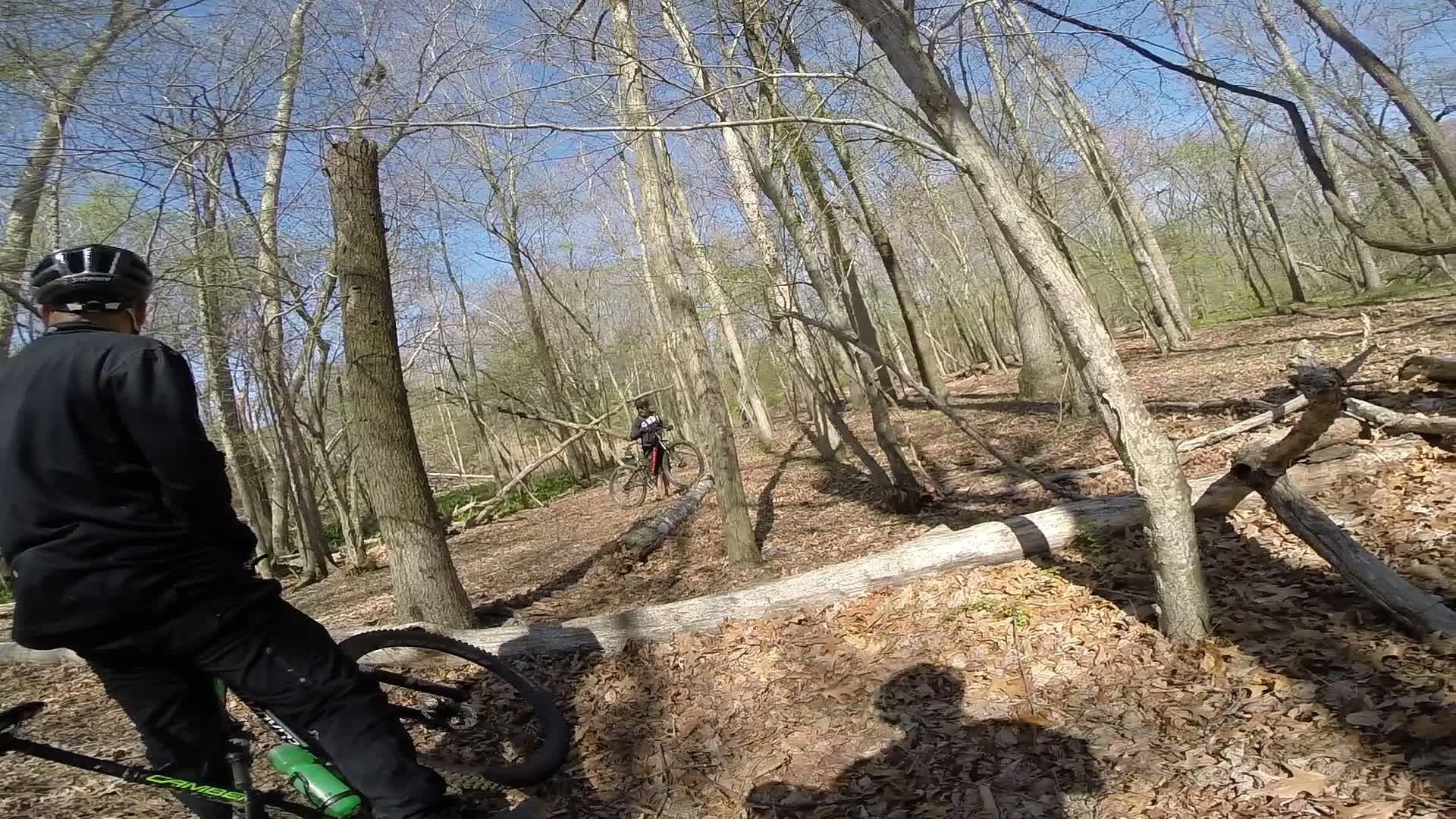 A person wearing a helmet and cycling gear stands beside a mountain bike on a wooded trail, while another cyclist rides in the background. The environment features bare trees and a forest floor covered in leaves and branches, under a clear blue sky. Allaire State Park mountain bike trail.