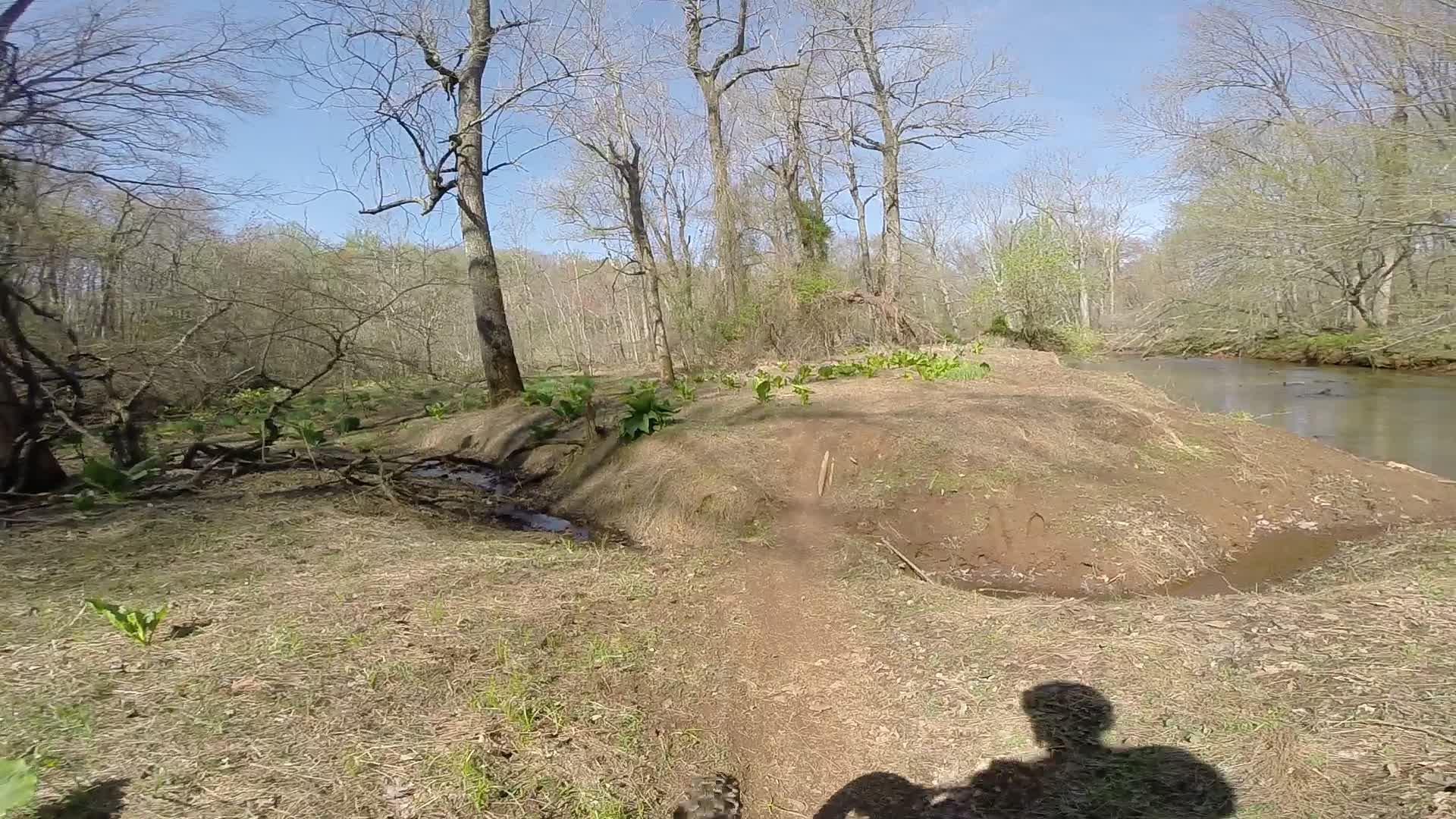 A scenic view of a forested area with bare trees, a small river, and patches of greenery along the banks. A dirt path winds through the landscape, while a shadow of a person is visible in the foreground. The sky is clear and blue, indicating a sunny day. Allaire State Park mountain bike trail.