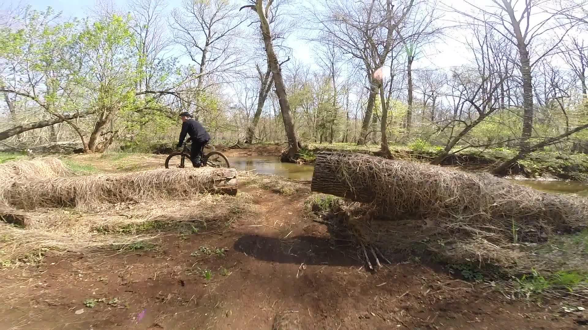 A mountain biker navigating a muddy trail covered with fallen logs in a wooded area, with a stream running alongside. The scene includes sparse trees with budding leaves, indicating early spring weather. Allaire State Park mountain bike trail.