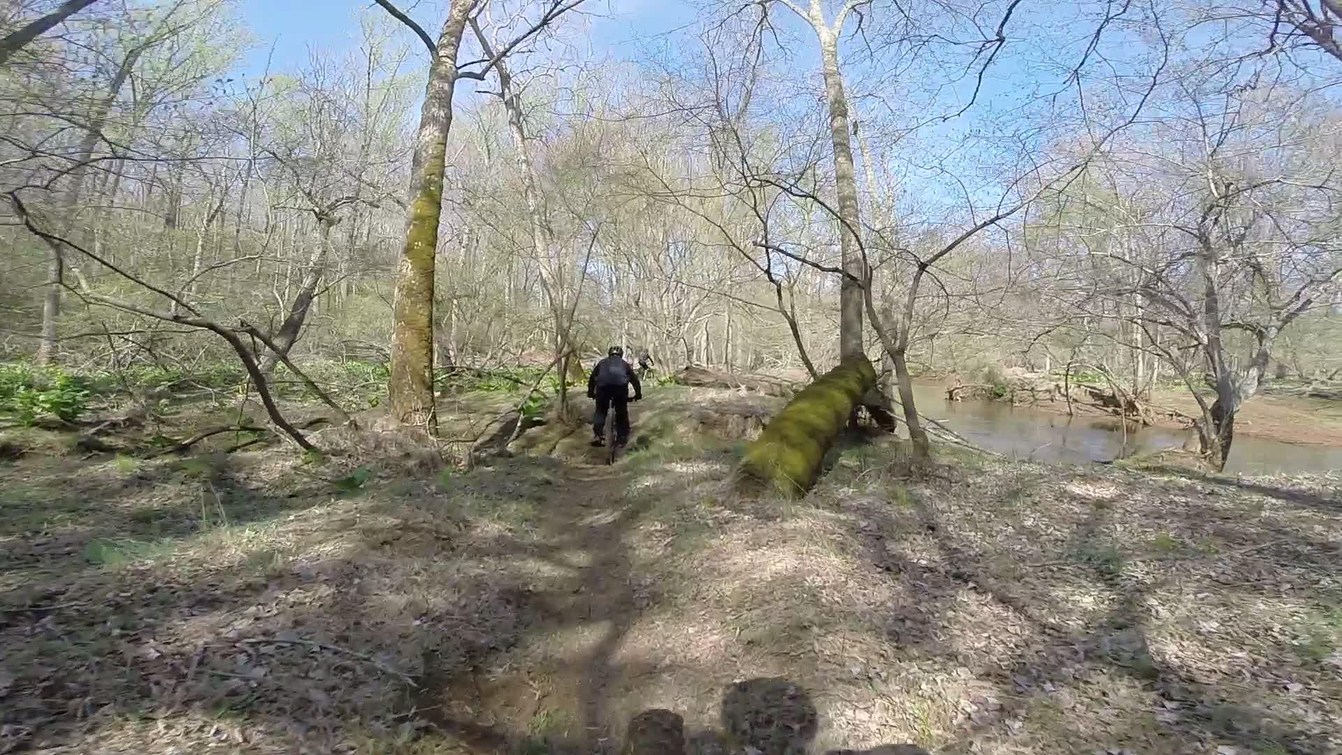 A cyclist riding along a narrow dirt trail in a forested area, surrounded by bare trees and underbrush, with a river visible to the right under a clear blue sky. Allaire State Park mountain bike trail.