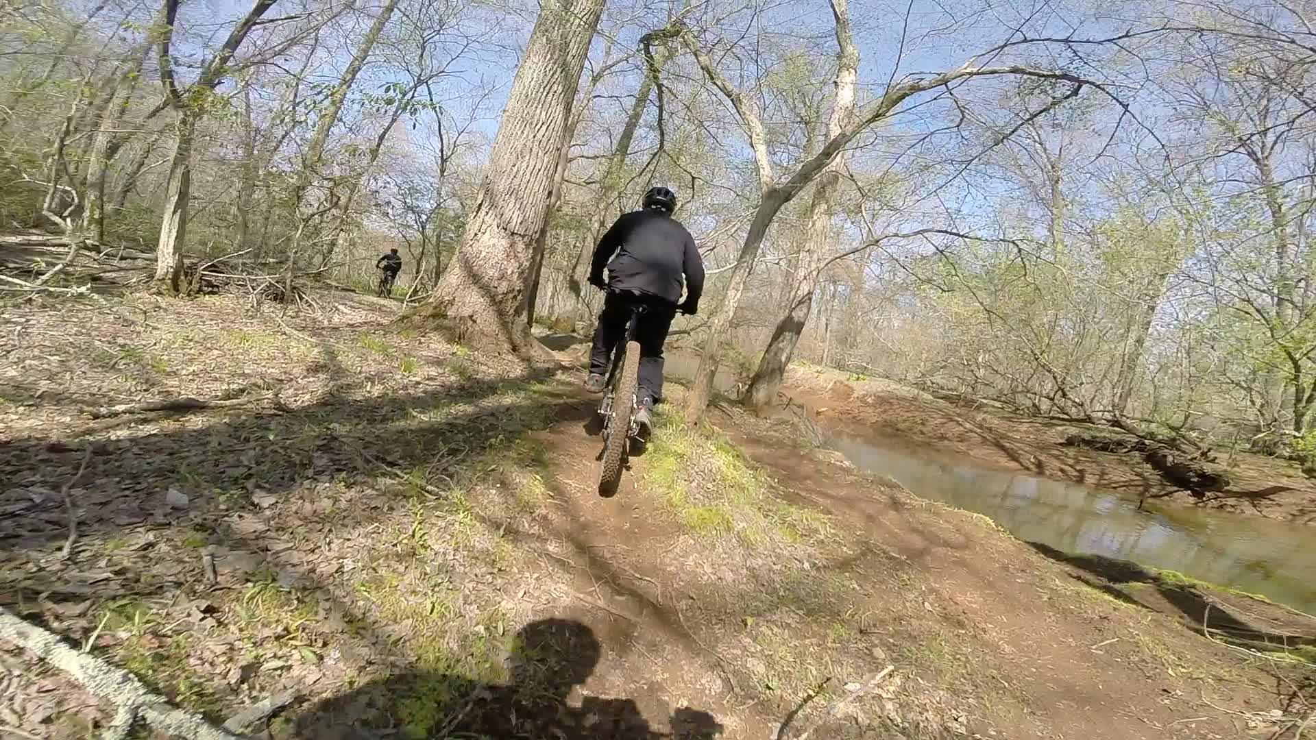 A mountain biker riding along a dirt path in a wooded area, with trees lining the trail and a shallow creek visible to the right. The scene is sunny, showcasing early spring foliage and a clear blue sky. Allaire State Park mountain bike trail.