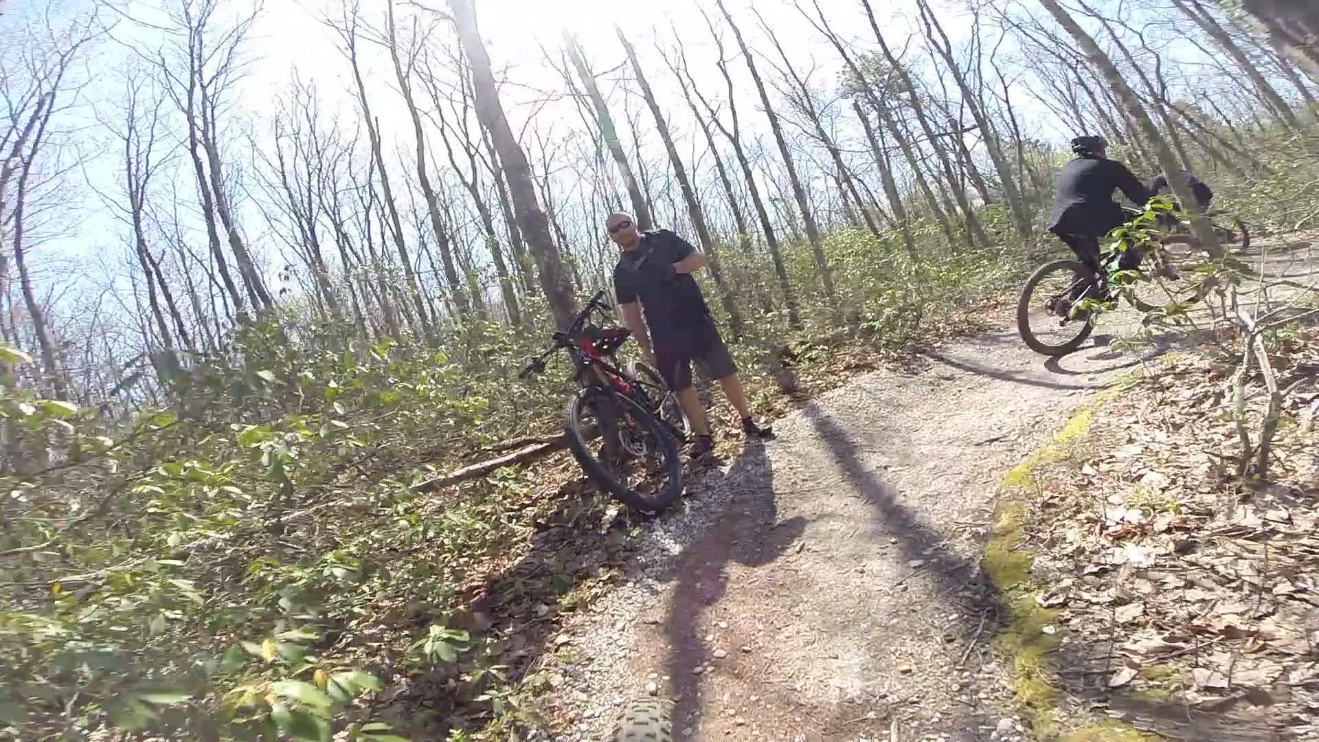 A mountain biker stands beside a bike on a dirt path in a wooded area, surrounded by leafless trees and green shrubbery, while another biker rides in the background. The sun shines brightly overhead, casting long shadows on the ground. Allaire State Park mountain bike trail.
