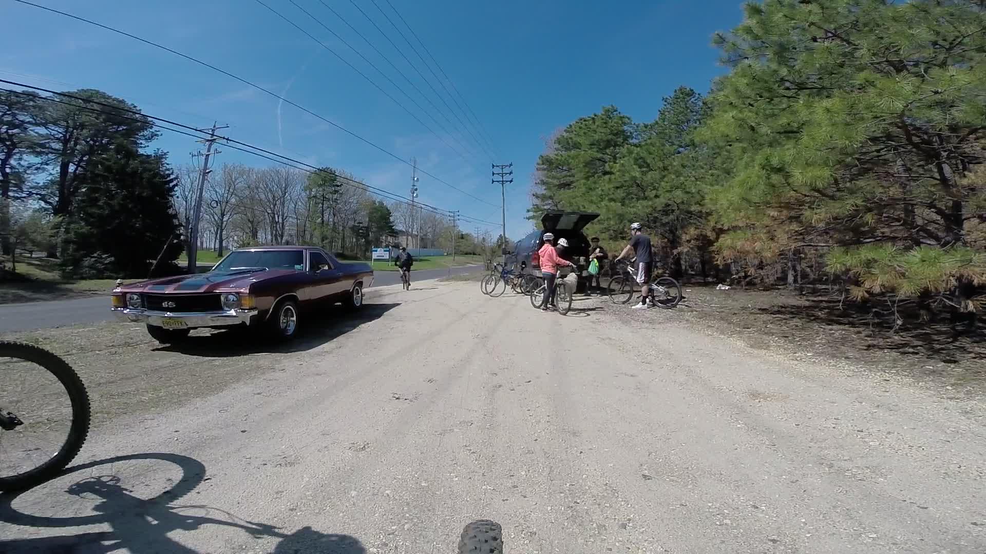 A group of cyclists is gathered near a dirt road, with some of them standing by their bicycles and others near a vehicle with its trunk open. A classic maroon car is parked nearby, and the area is surrounded by green trees under a clear blue sky. Allaire State Park mountain bike trail.
