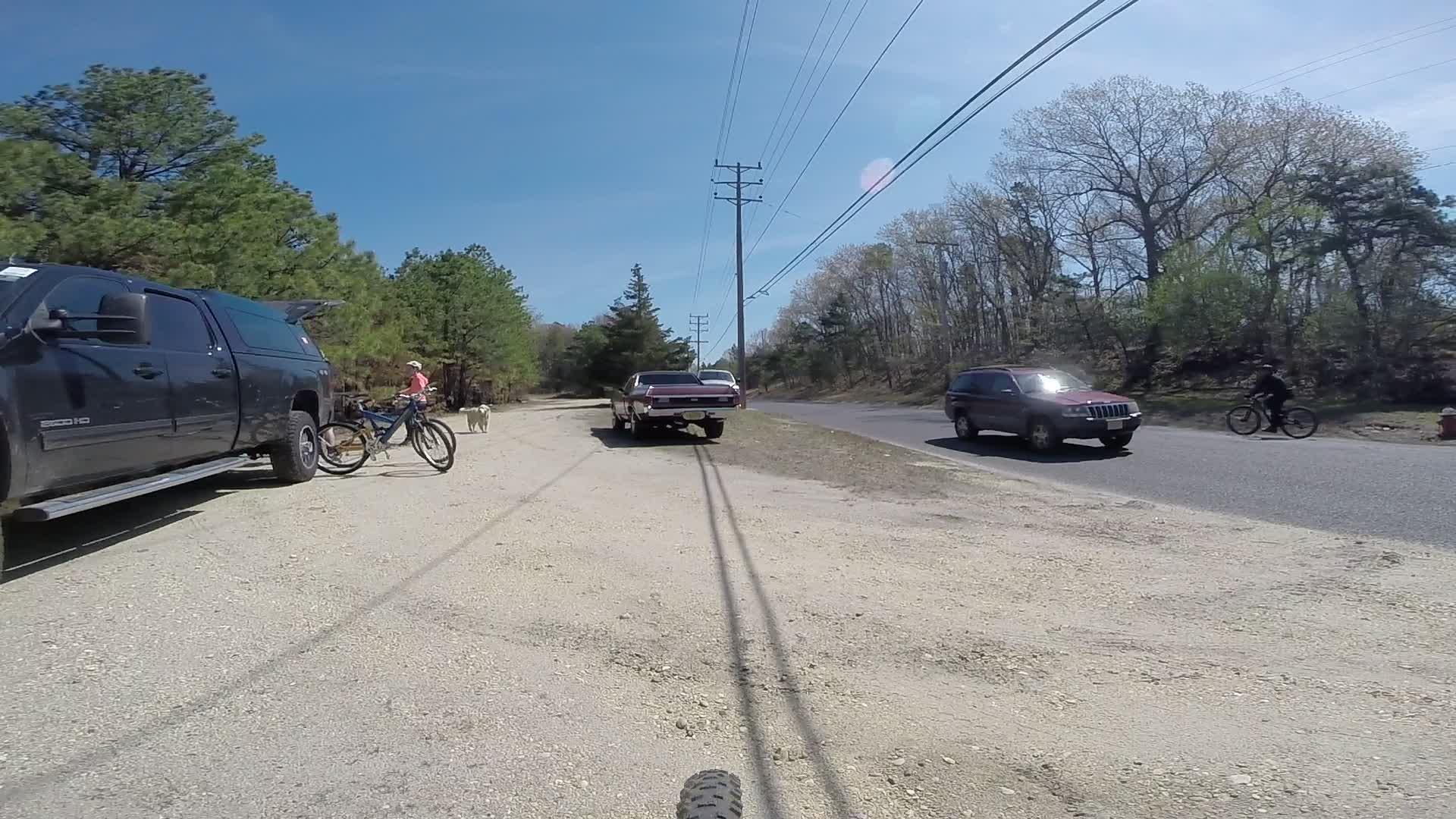 A gravel road scene on a sunny day, featuring a parked black pickup truck and a vintage car on the right. A cyclist rides past on the road, while a person in a pink shirt stands next to a bicycle near the truck. A dog is also visible, and trees line the background with power lines overhead. Allaire State Park mountain bike trail.