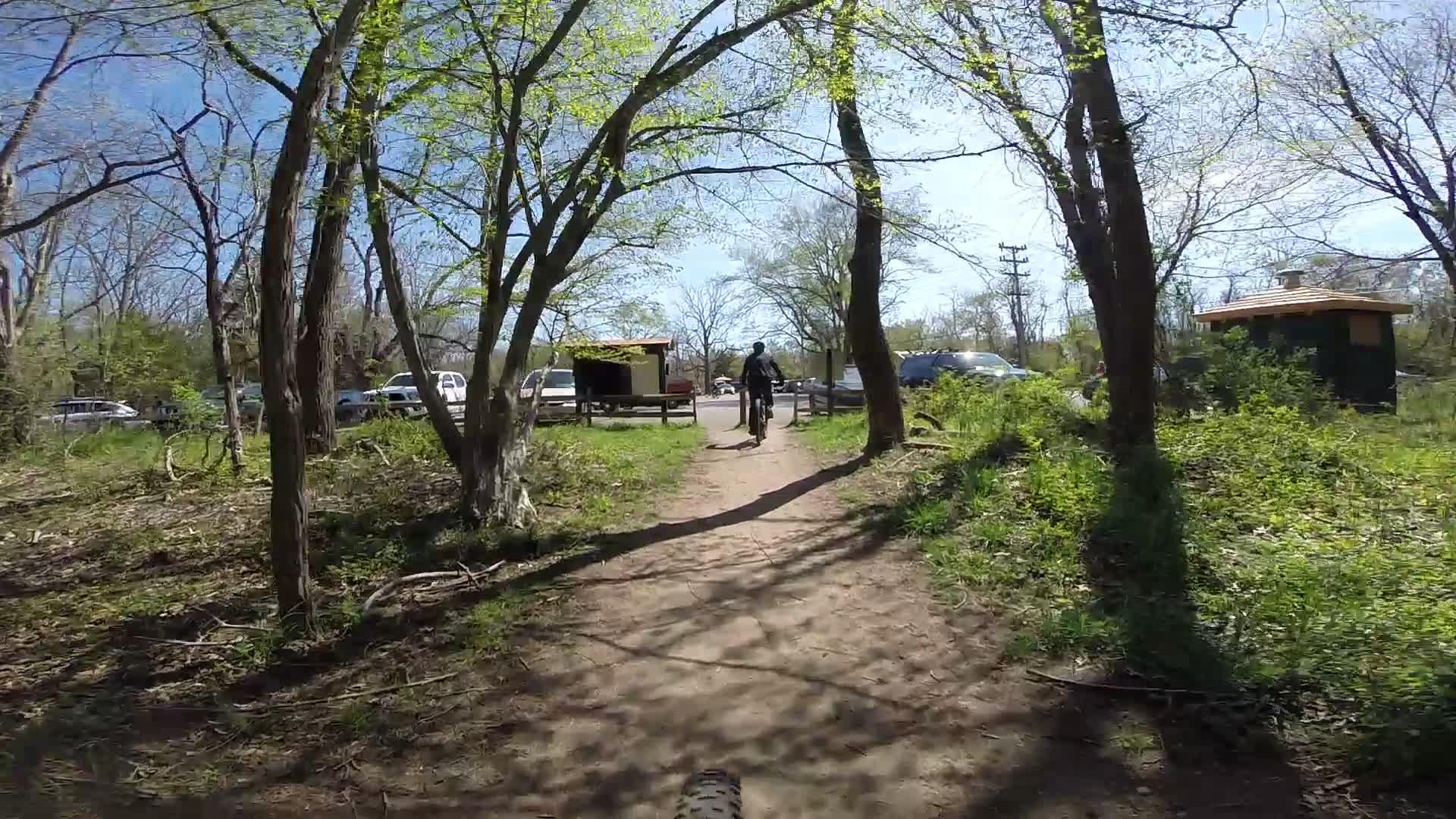 A dirt path winding through a wooded area with green leaves on the trees. A cyclist is riding away from the camera, heading towards a parking area with several vehicles. There is a small structure nearby, partially obscured by trees, and the sky is clear with bright sunshine. Allaire State Park mountain bike trail.