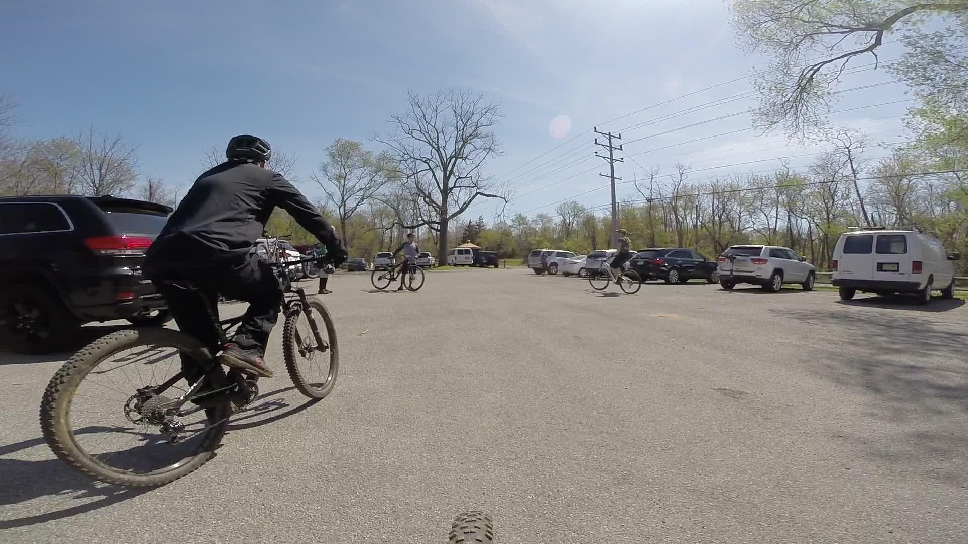 A group of mountain bikers in a parking lot with several parked cars. The scene is set on a sunny day, with trees in the background and power lines visible. One cyclist is seen riding away from the camera, while others prepare to start their ride. Allaire State Park mountain bike trail.