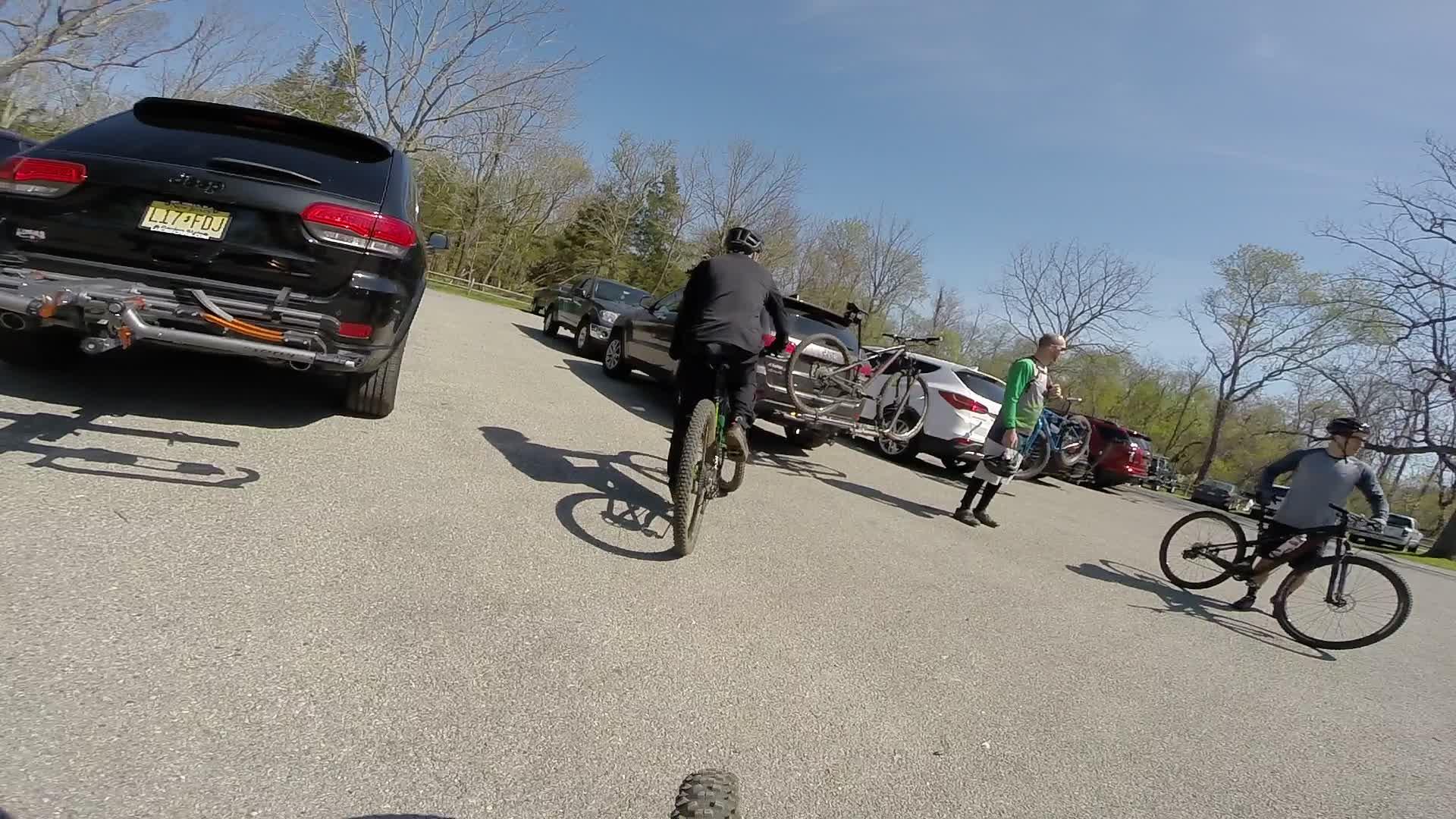 A group of mountain bikers preparing for a ride in a parking lot on a sunny day. Several bicycles are mounted on vehicle racks, and two riders are seen discussing while another one begins to ride away. Vehicles are parked in the background, with trees in the distance signaling an outdoor recreational area. Allaire State Park mountain bike trail.