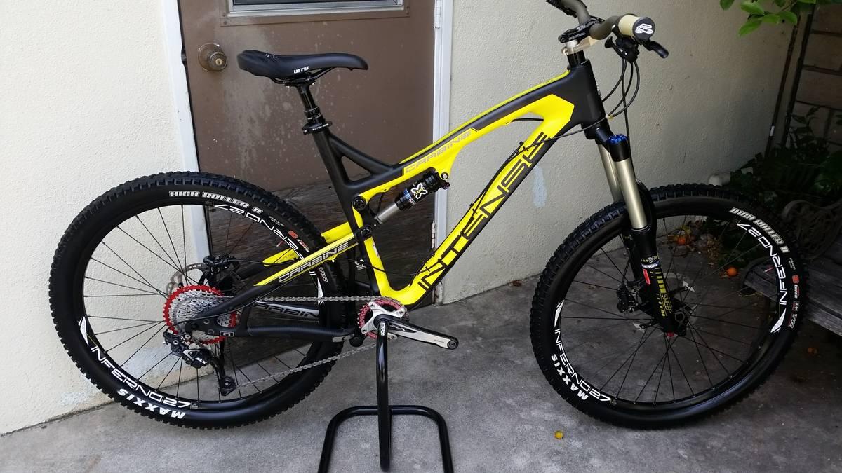 Intense Carbine 275: A vibrant yellow and black mountain bike is displayed on a stand against a light-colored wall. The bike features chunky tires, a suspension system, and a sleek frame design. It is positioned near a door, with some greenery visible in the background.