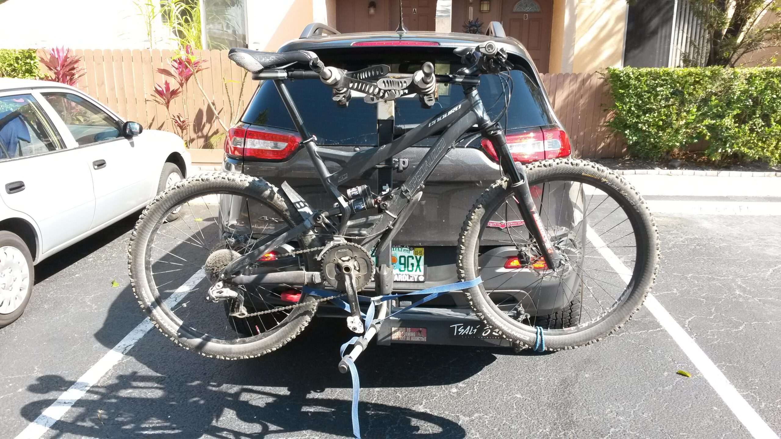 Specialized Enduro Comp: A black mountain bike is mounted on the back of an SUV, secured with a blue strap. The bike has dirty tires, indicating recent use, and the SUV's rear license plate is partially visible. In the background, another white car is parked nearby, along with plants and a wooden fence.