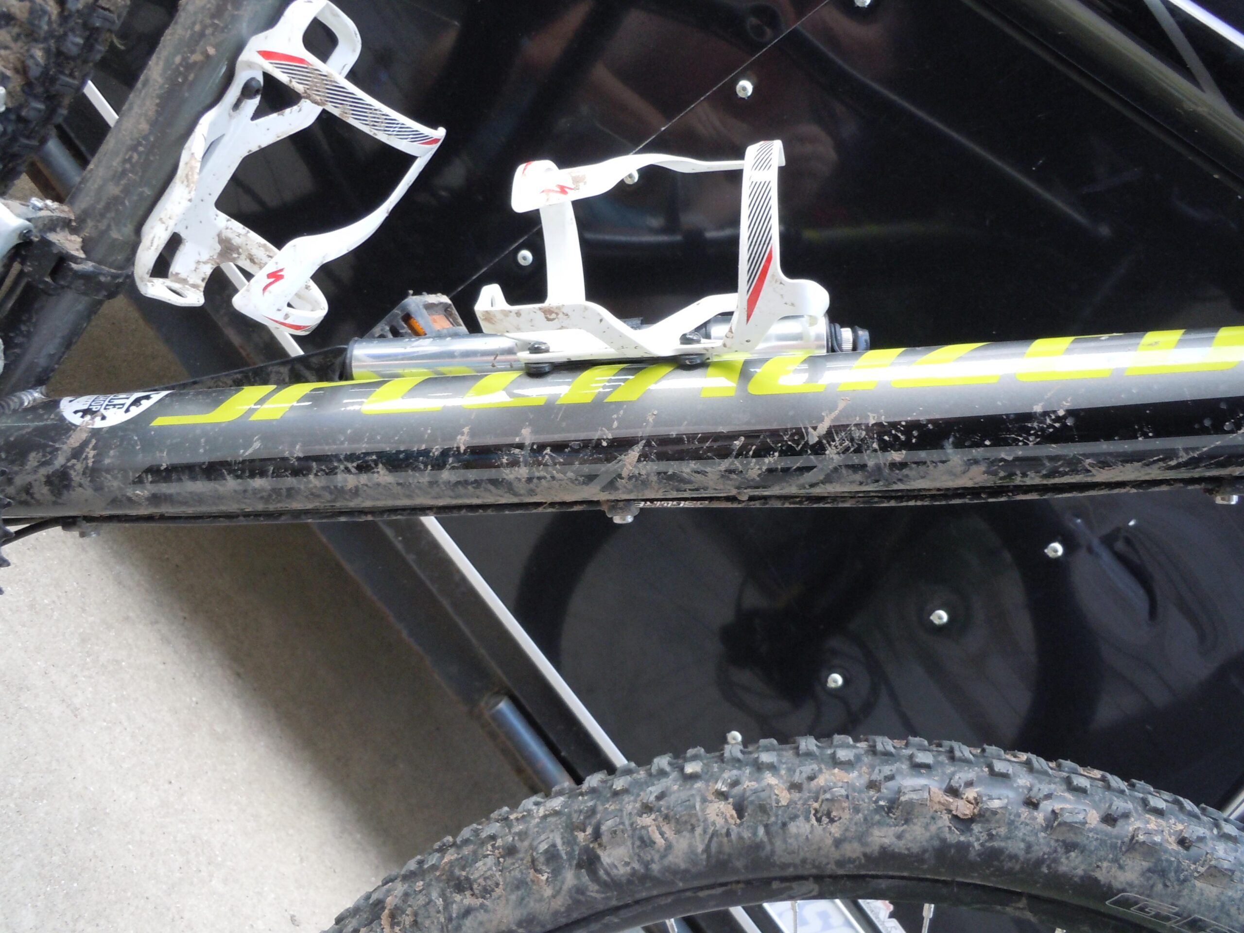 Specialized Rockhopper 29: A close-up view of a mountain bike frame with a yellow logo, showcasing dirt and grime from recent use. The bike features white pedal clips and a visible gear mechanism, with a blurred black background suggesting storage or transport.