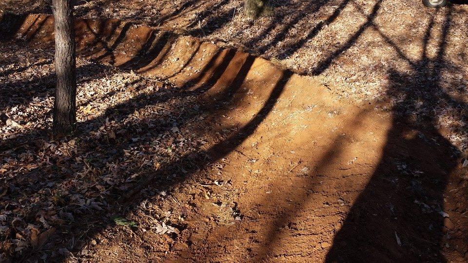A dirt bike trail in a wooded area, showing a series of dirt berms and shadows from nearby trees. The ground is covered with dry leaves, and the soil appears rich and brown. Mountain Laurel Trails mountain bike trail.