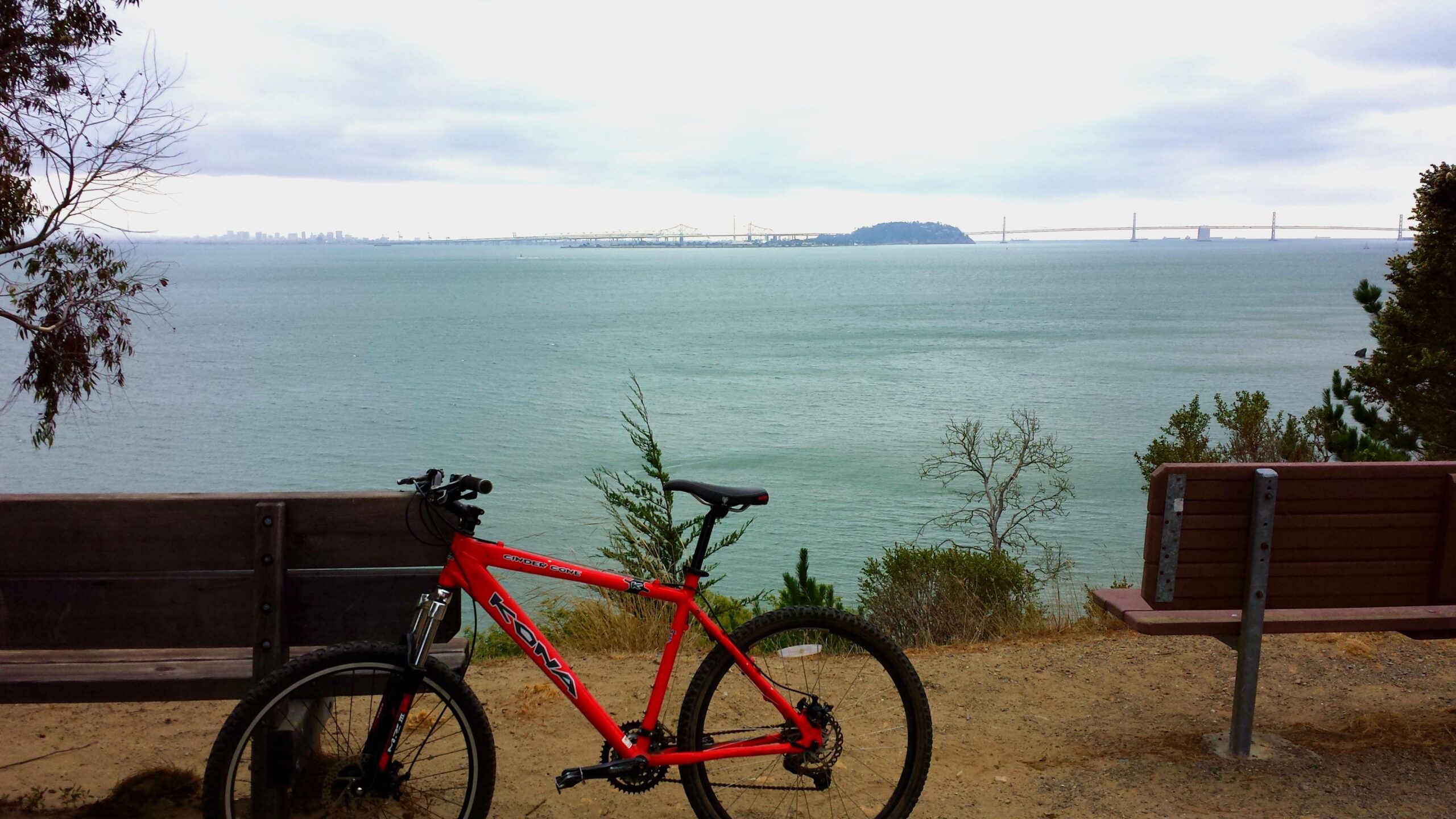 Kona Cinder Cone: A bright red mountain bike is parked beside a wooden bench overlooking a calm body of water, with distant views of a bridge and city skyline under a cloudy sky. Green foliage surrounds the scene, creating a serene outdoor setting.
