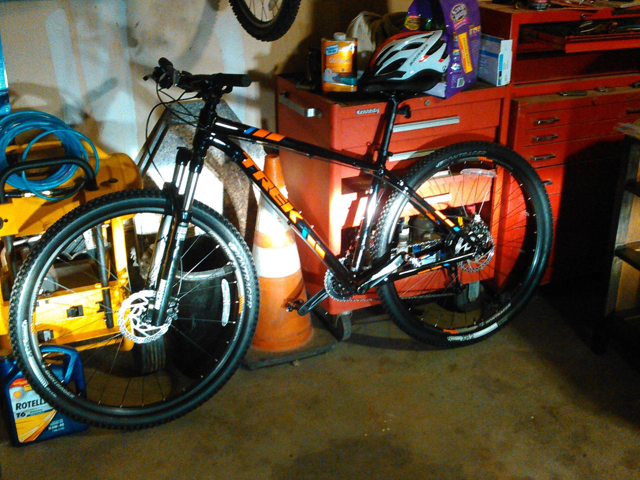 Trek Trek x caliber 8: A black mountain bike with orange and blue accents leaning against a red tool chest in a garage. A white helmet is placed on top of the bike, and various tools and supplies are visible in the background, including a stack of boxes and a traffic cone. The garage floor is concrete, and the lighting creates a warm, slightly shadowy atmosphere.