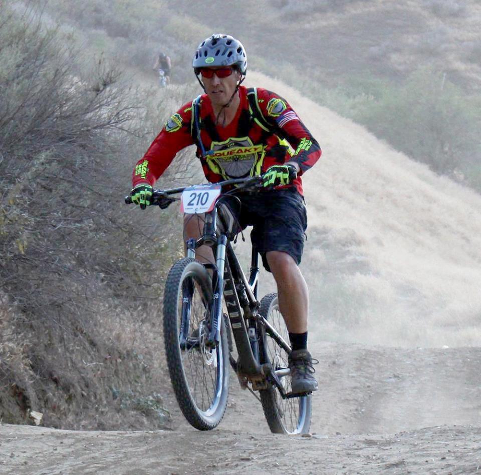 Mountain biker wearing a helmet and sunglasses, dressed in a red and yellow jersey, racing down a dusty trail with shrubbery on the side. The rider has a race number "210" displayed on their bike. A blurred figure can be seen in the background on a hill. Castaic Lake Pro Uphill mountain bike trail.