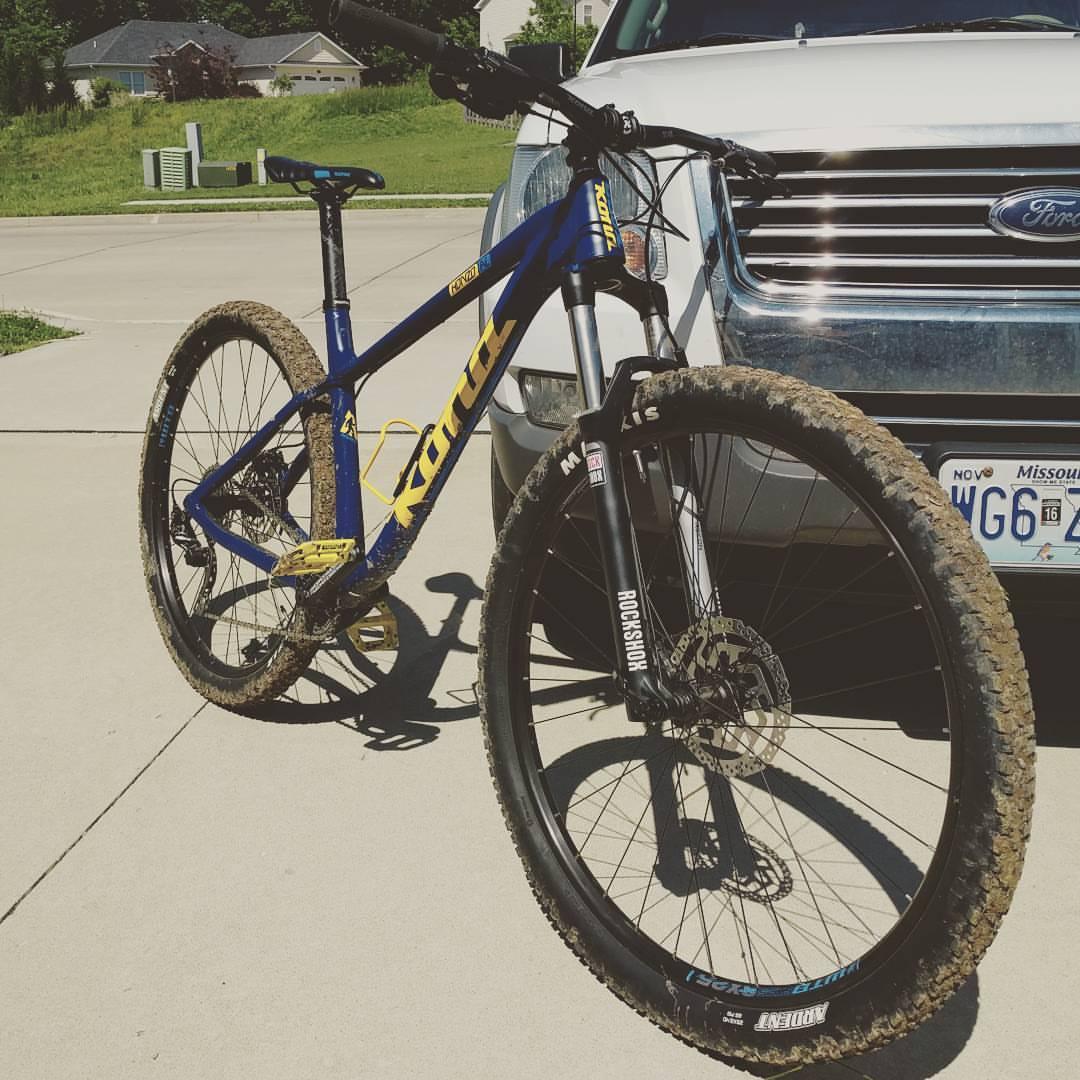 Kona Honzo: A blue and yellow mountain bike is parked on a concrete surface next to a silver SUV. The bike has mud on its tires, indicating recent use. In the background, there are grassy areas and residential buildings.