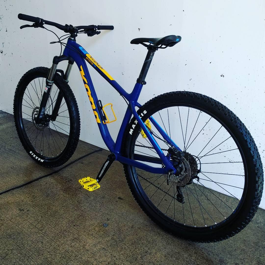 Kona Honzo: A blue and yellow mountain bike is positioned against a plain white wall. The bike features thick, knobby tires marked with "Maxxis" on the sidewalls and distinctive yellow flat pedals. It has a sturdy frame and front suspension, showcasing a modern design ideal for off-road cycling. The background is a concrete floor with minimal texture.