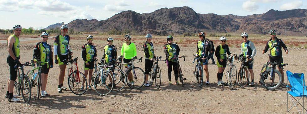 A group of twelve cyclists wearing matching green and black cycling jerseys and helmets poses next to their bicycles on a rocky, open landscape with mountains in the background. The scene is set under a partly cloudy sky, and one cyclist is sitting on a blue folding chair nearby.