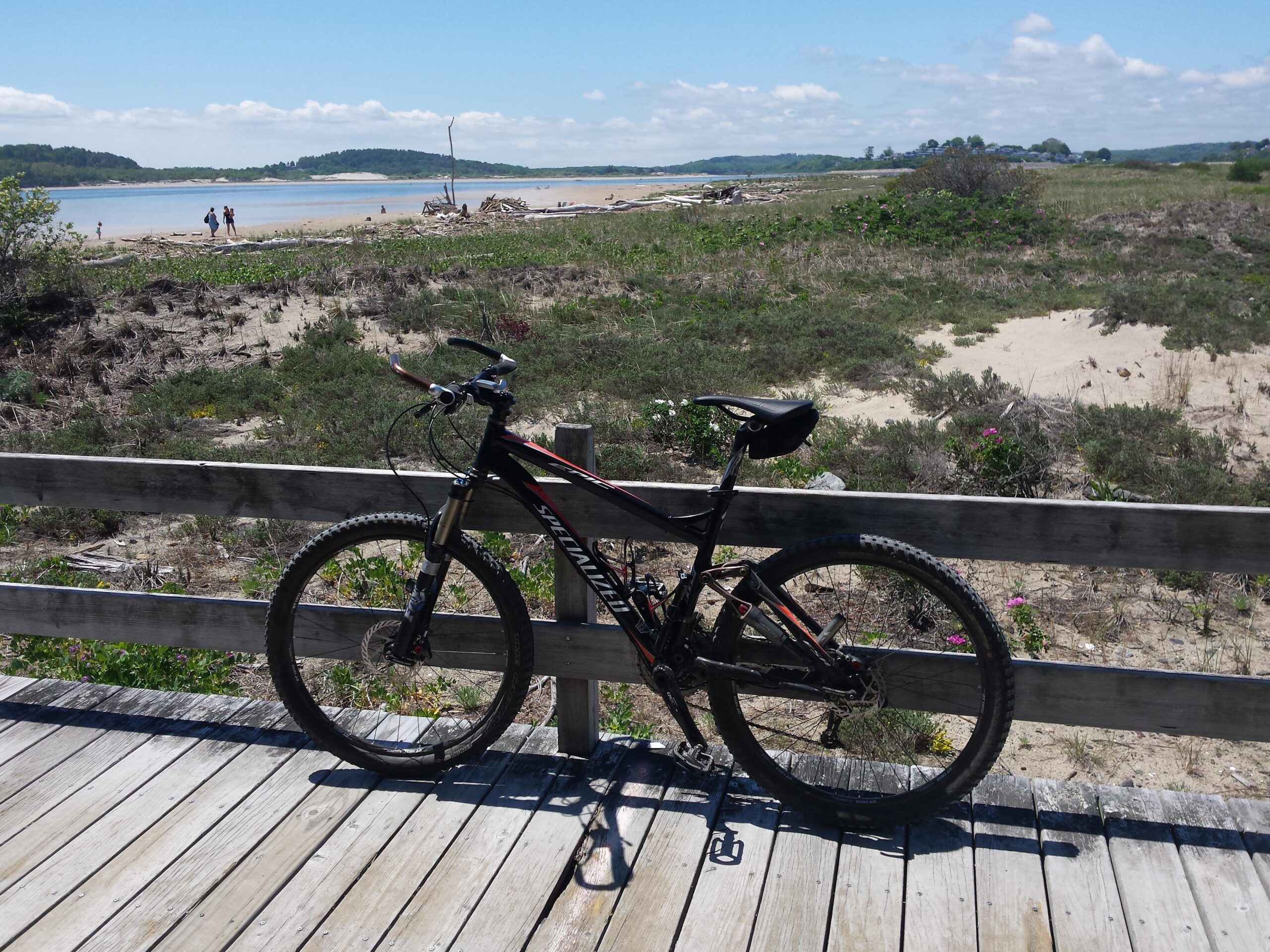 Specialized Epic Expert: A mountain bike parked on a wooden boardwalk, with a scenic view of a sandy beach, blue water, and distant hills under a partly cloudy sky. In the background, people can be seen walking along the shore, surrounded by lush greenery and a variety of plants.