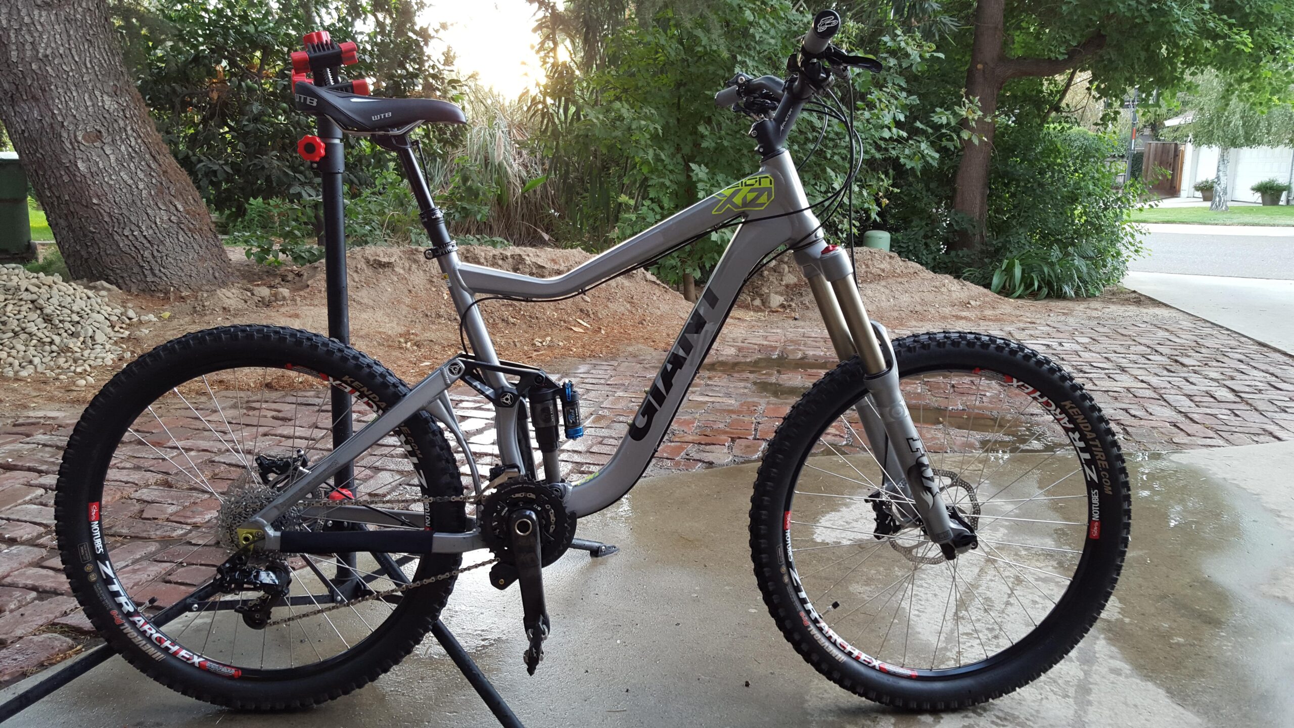 Giant Reign: A mountain bike with a gray frame and colorful accents is positioned on a bike stand. The bike features wide tires designed for off-road terrain, along with a detailed drivetrain. The background shows lush greenery and a driveway, indicating an outdoor setting. The ground is wet, suggesting recent cleaning or rain.