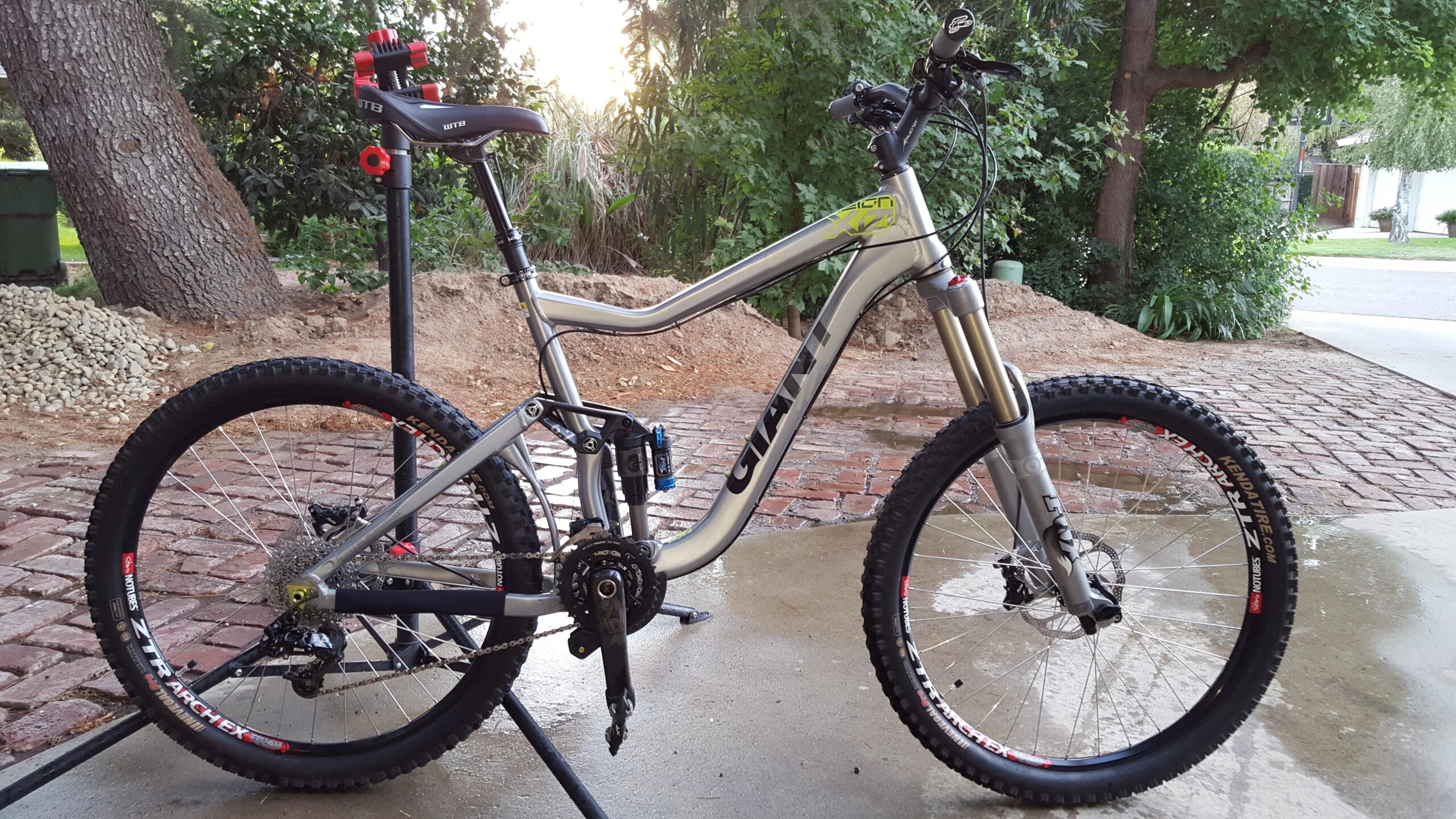 Giant Reign: A silver mountain bike with black accents, prominently featuring the "Giant" brand, is positioned on a concrete surface. The bike is equipped with knobby tires, a suspension system, and a visible gear mechanism. Behind it, there is a tree and shrubs, indicating an outdoor setting, while a brick pathway and a green trash bin are also visible in the background.