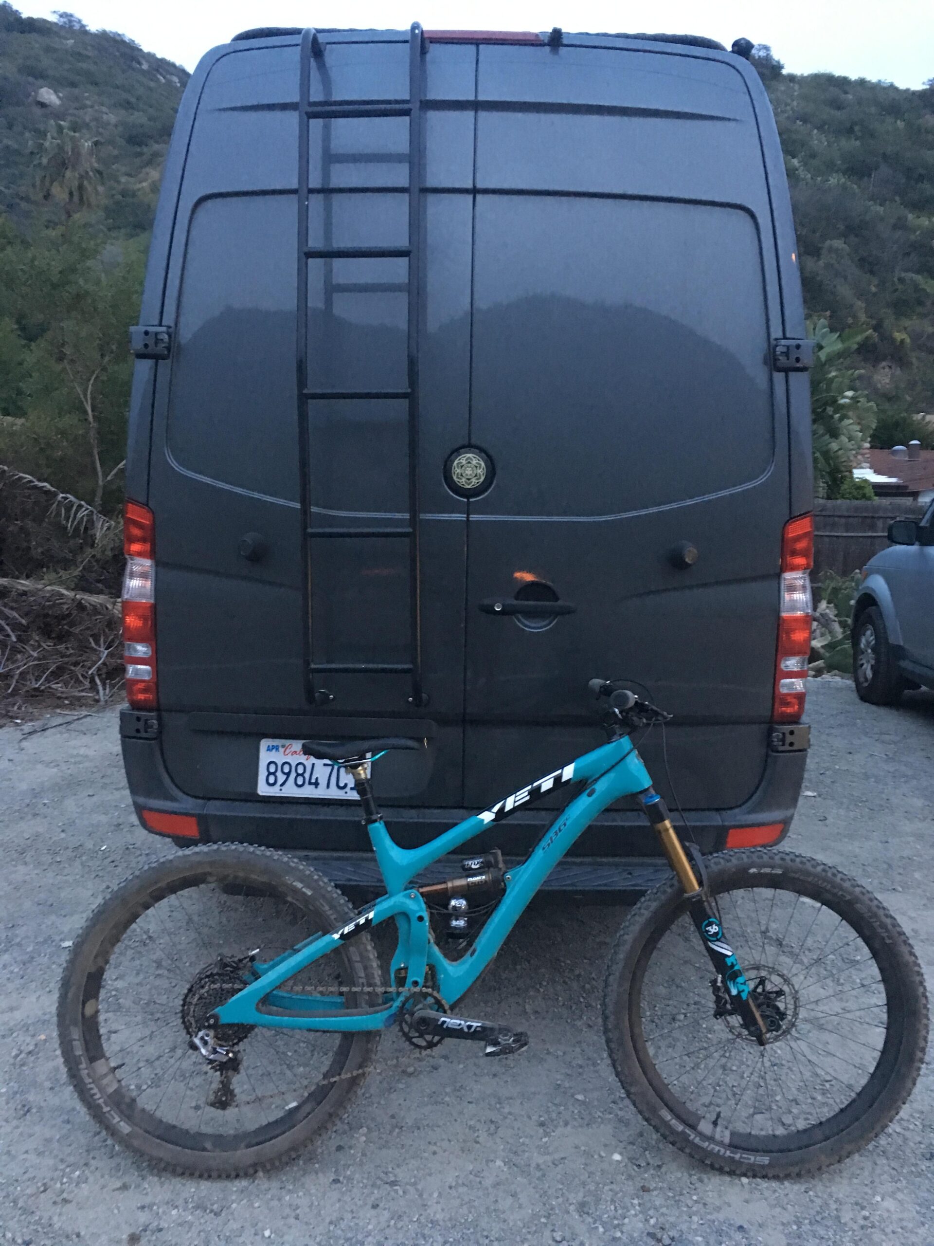 Yeti SB6: Alt tag: A teal mountain bike leaning against the back of a dark gray van, which features a ladder attached to its side. The scene is set in a gravel area with greenery and hills in the background, taken during twilight.