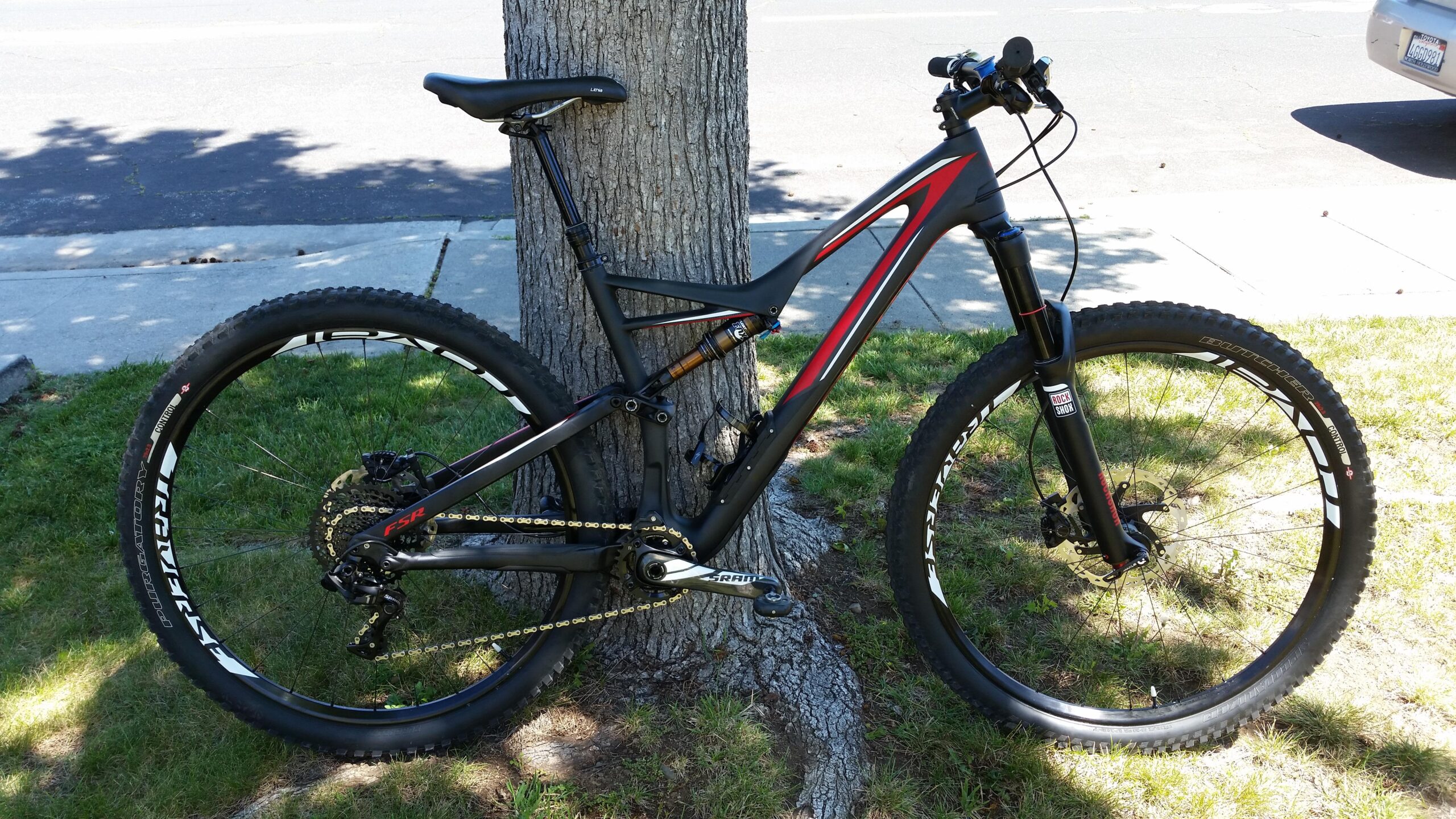 Specialized Stumpjumper FSR Expert 29: A mountain bike leaning against a tree on a grassy area, featuring a black frame with red accents, thick tires, and a suspension system. The background shows a sidewalk and a road, with sunlight filtering through the leaves.