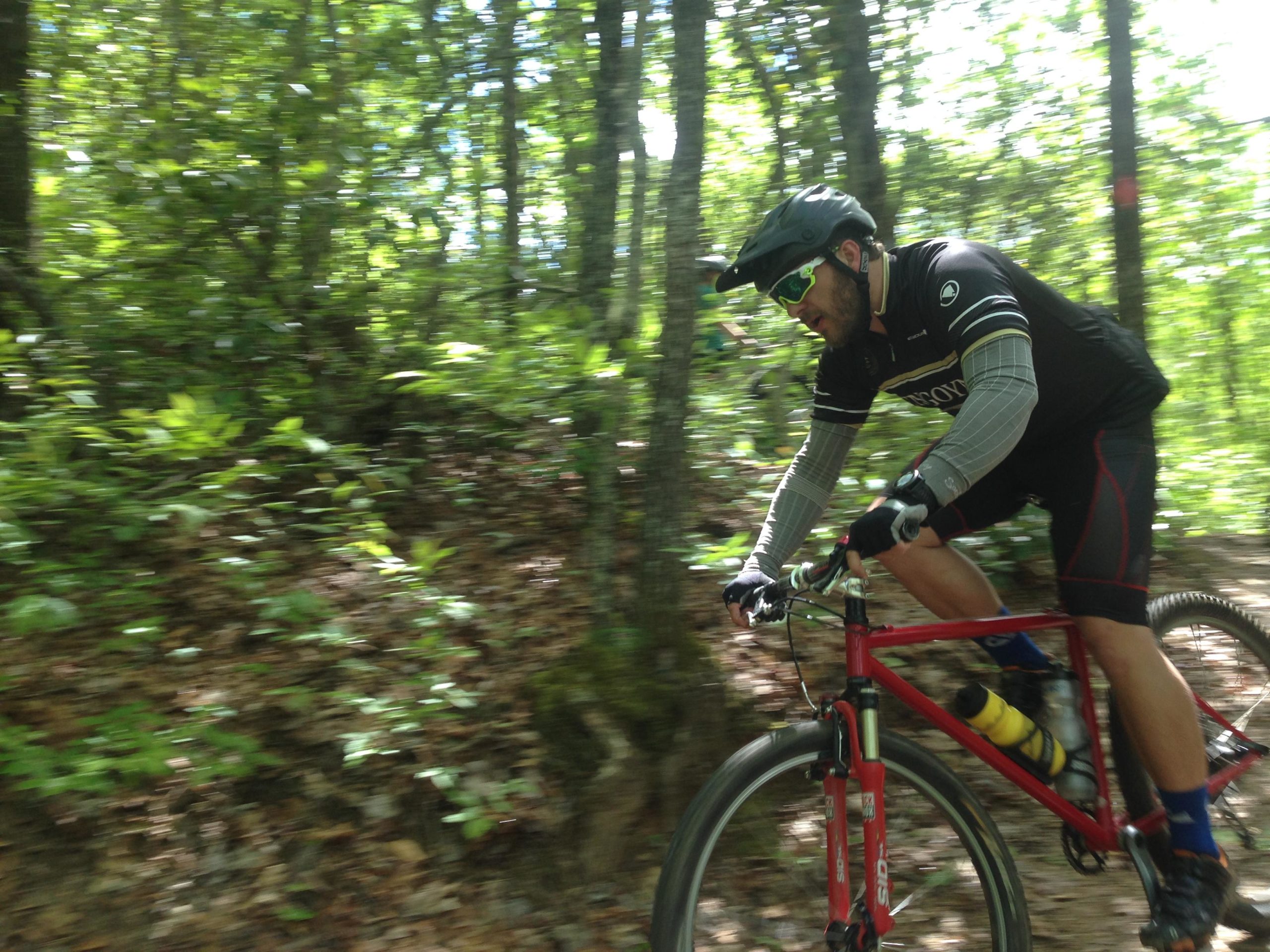 A mountain biker navigating a dirt trail surrounded by lush green trees and sunlight filtering through the leaves. The rider is wearing a helmet and sunglasses, focused on the path ahead. The motion blur captures the feeling of speed and adventure. Bracken Preserve mountain bike trail.