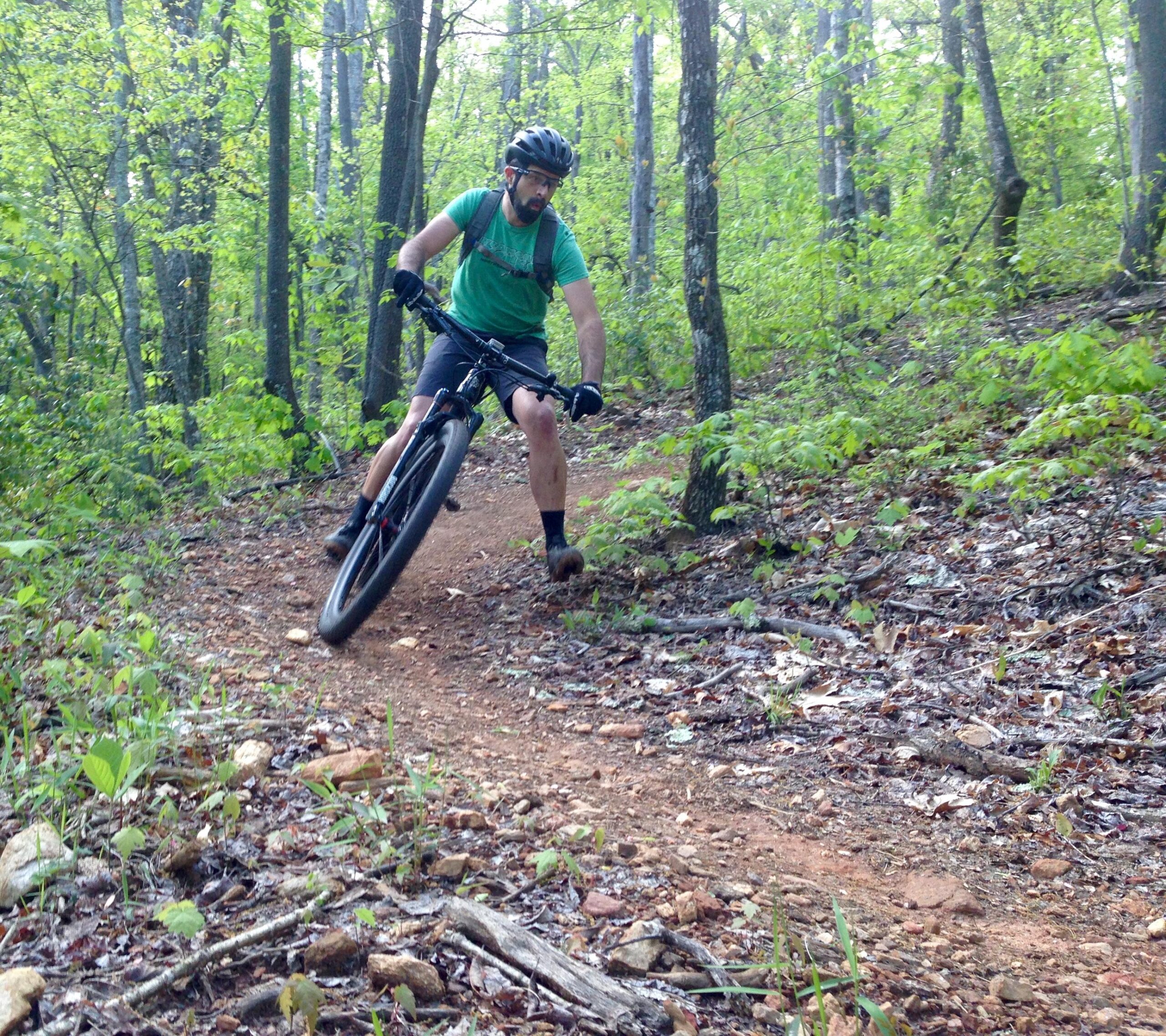 A cyclist navigating a winding dirt trail in a lush green forest, leaning into a turn on a mountain bike. The rider is wearing a helmet, a green shirt, and shorts, with trees and foliage in the background. Bracken Preserve mountain bike trail.