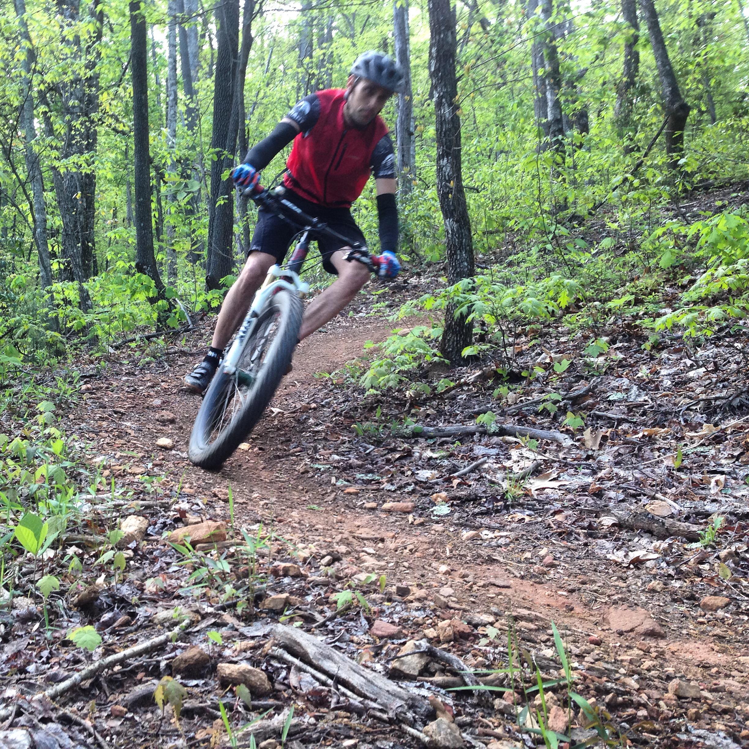 A cyclist in a red vest and black cycling gear is riding a mountain bike on a winding dirt trail surrounded by lush green trees and underbrush. The bike is leaning into a turn, creating a dynamic and action-packed scene in a forested area. Bracken Preserve mountain bike trail.