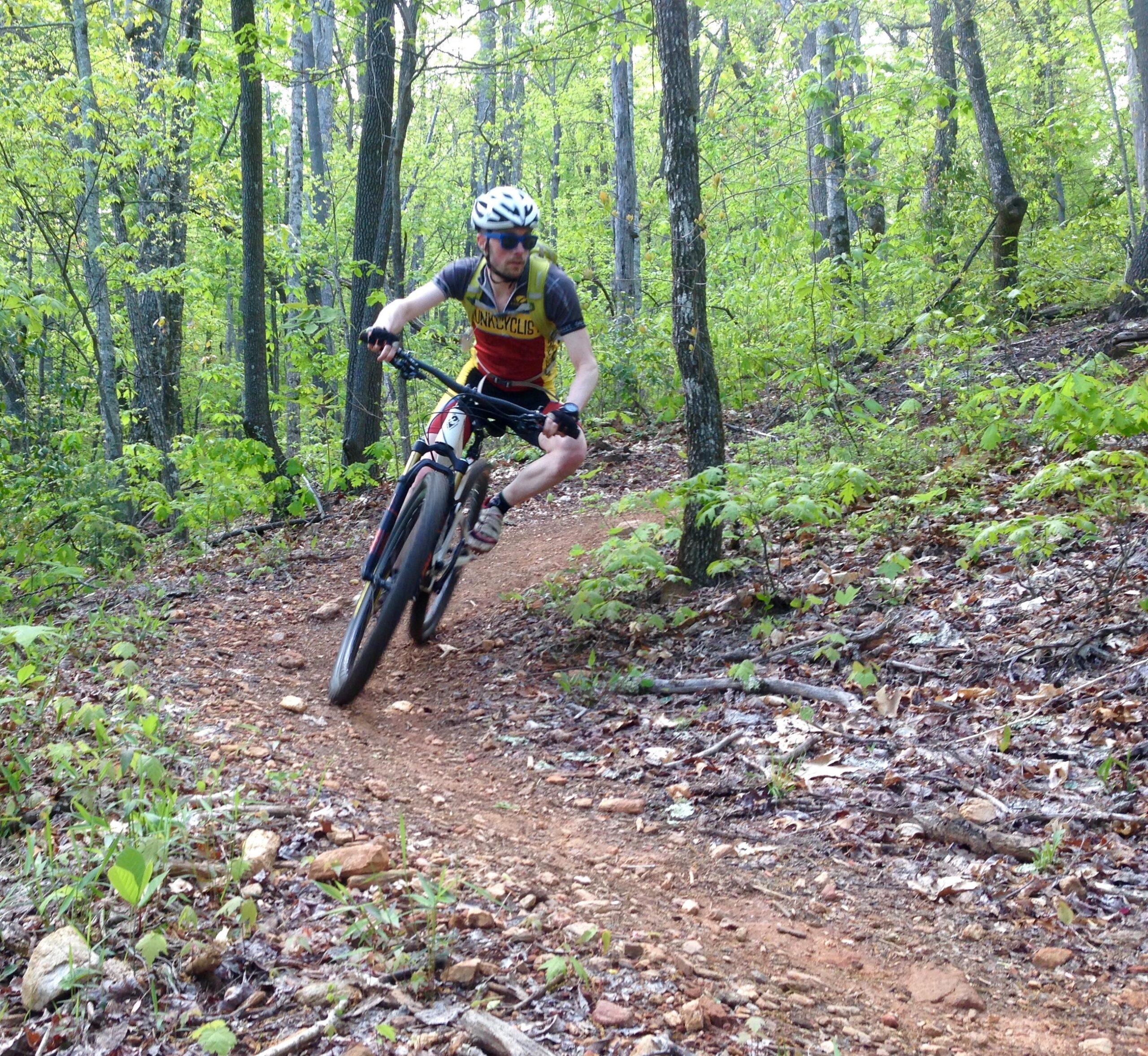 A mountain biker navigating a winding dirt trail through a lush green forest, wearing a helmet and sunglasses, and dressed in a colorful jersey and shorts. Bracken Preserve mountain bike trail.