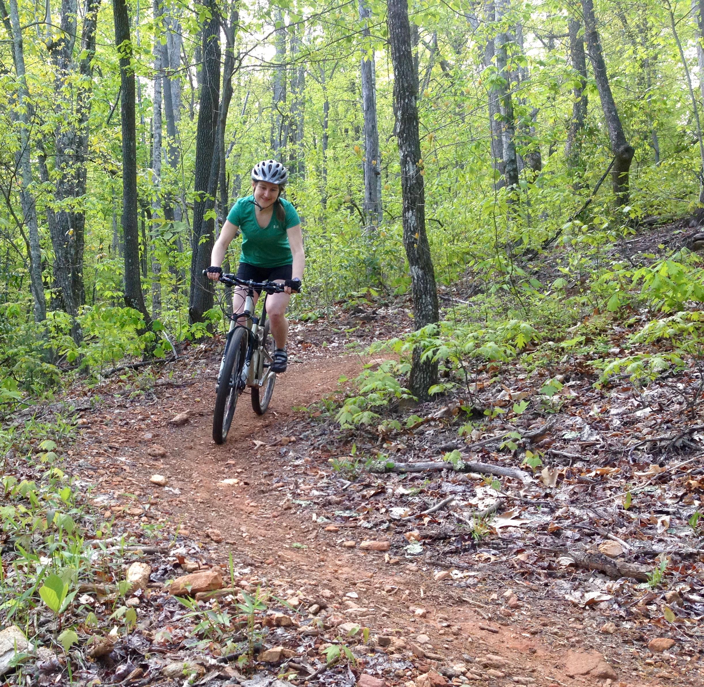 A person riding a mountain bike on a dirt trail surrounded by lush green trees and foliage, wearing a helmet and a green shirt. Bracken Preserve mountain bike trail.