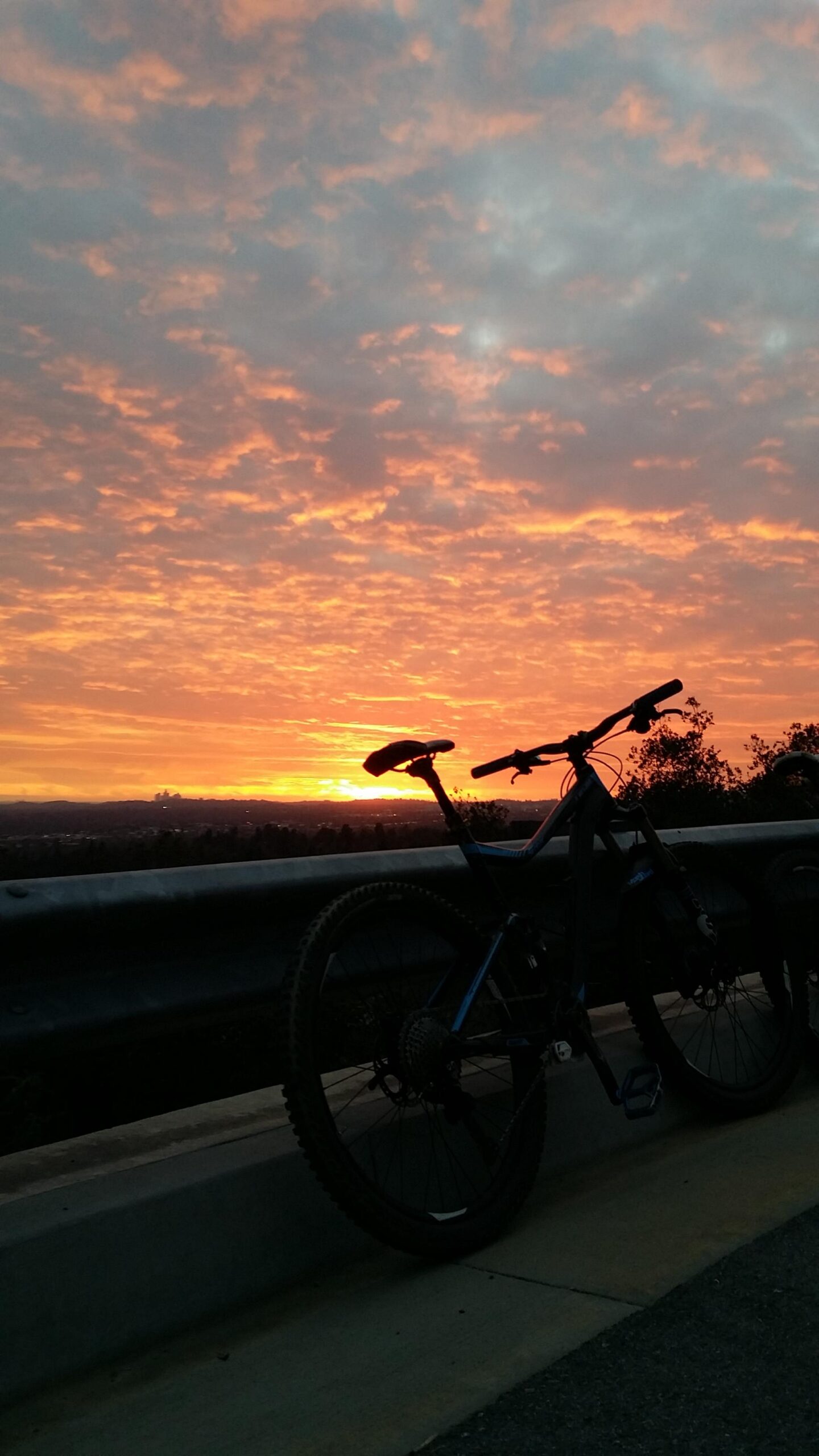 Giant Giant Trance 27.5 2: A silhouette of a mountain bike leaning against a guardrail at sunset, with vibrant orange and pink clouds in the sky and a distant horizon.