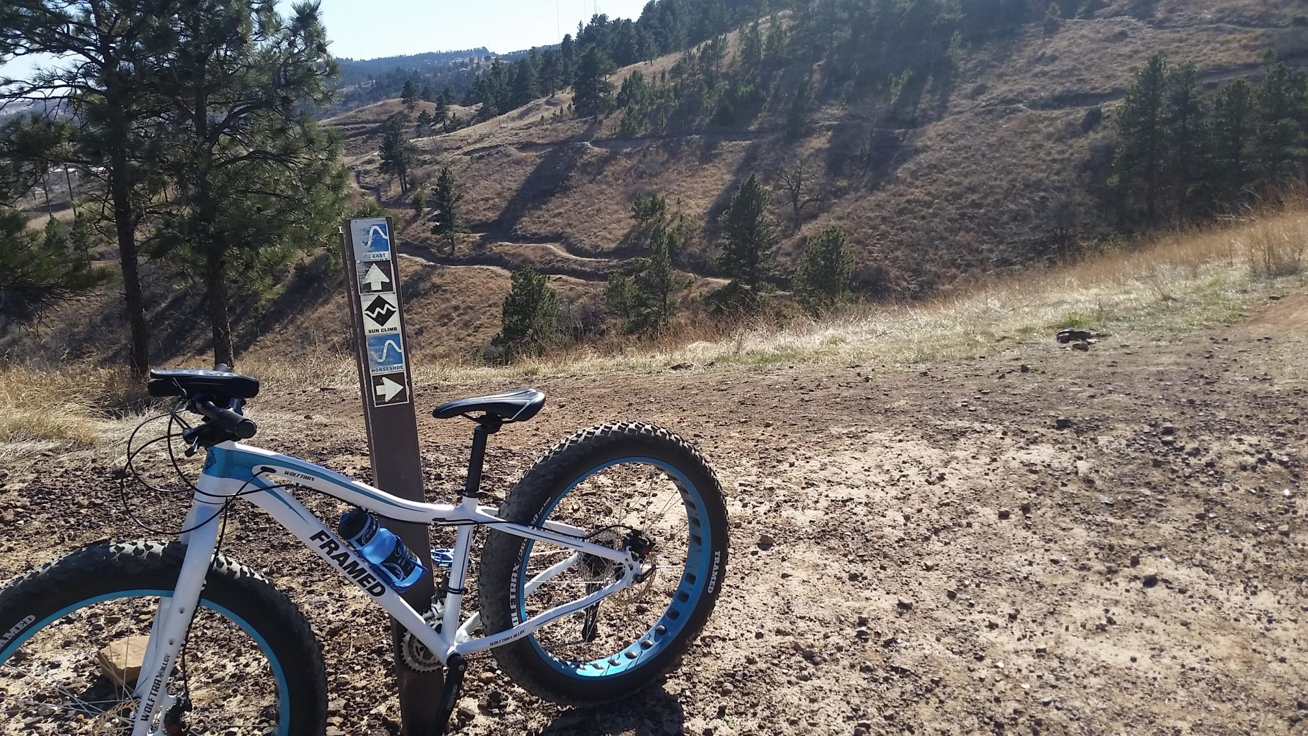 A mountain bike parked next to a trail sign on a dirt path, with rolling hills and trees in the background under a clear blue sky. The bike features fat tires and a water bottle attached to the frame. The trail sign displays markers for various biking routes. HLMP mountain bike trail.