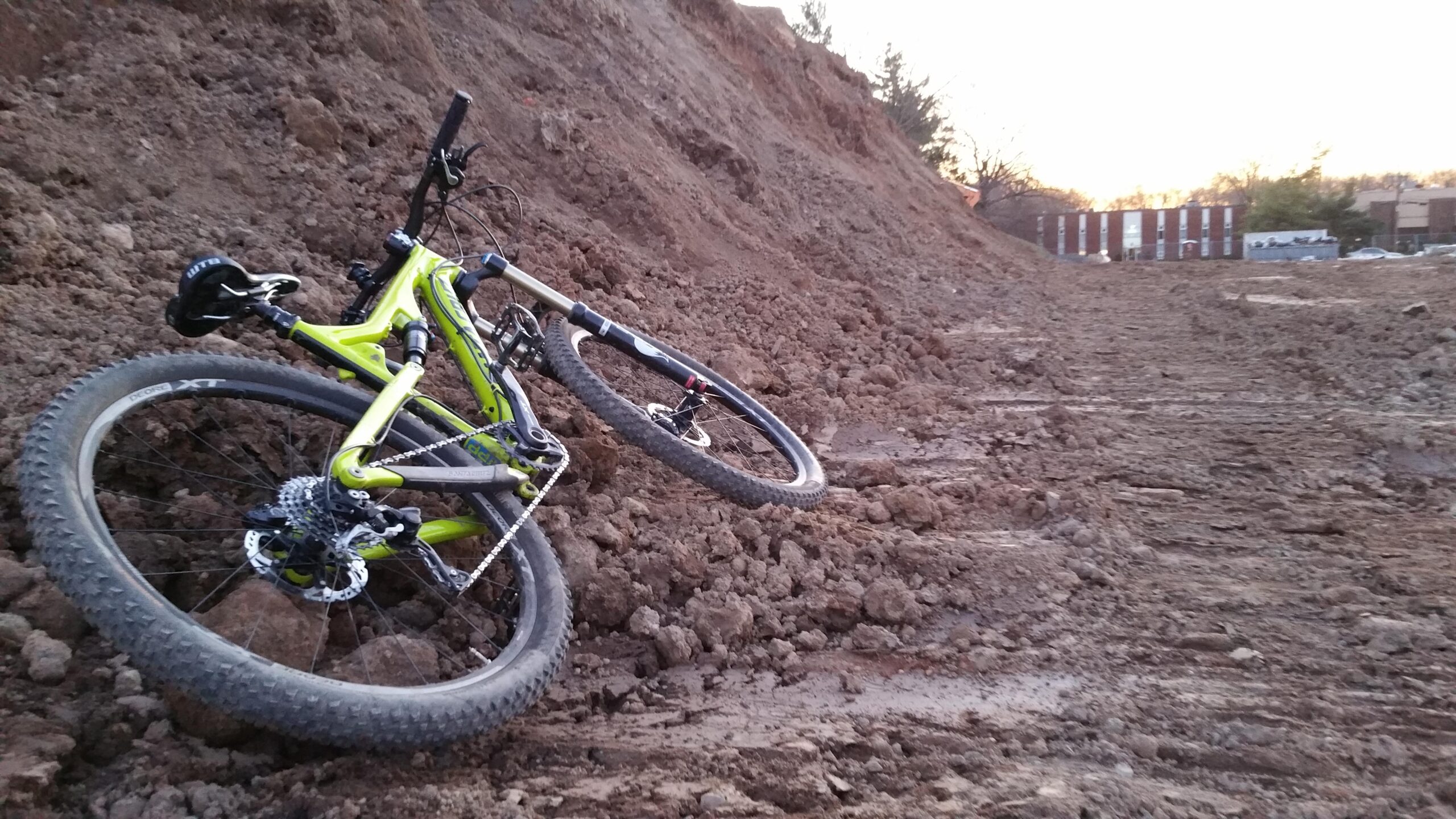 Santa Cruz Bronson C: A mountain bike leaning against a dirt embankment, surrounded by freshly turned soil, with a sunset in the background. The bike features vibrant green and black colors and is positioned on rough terrain, indicating a rugged outdoor setting.