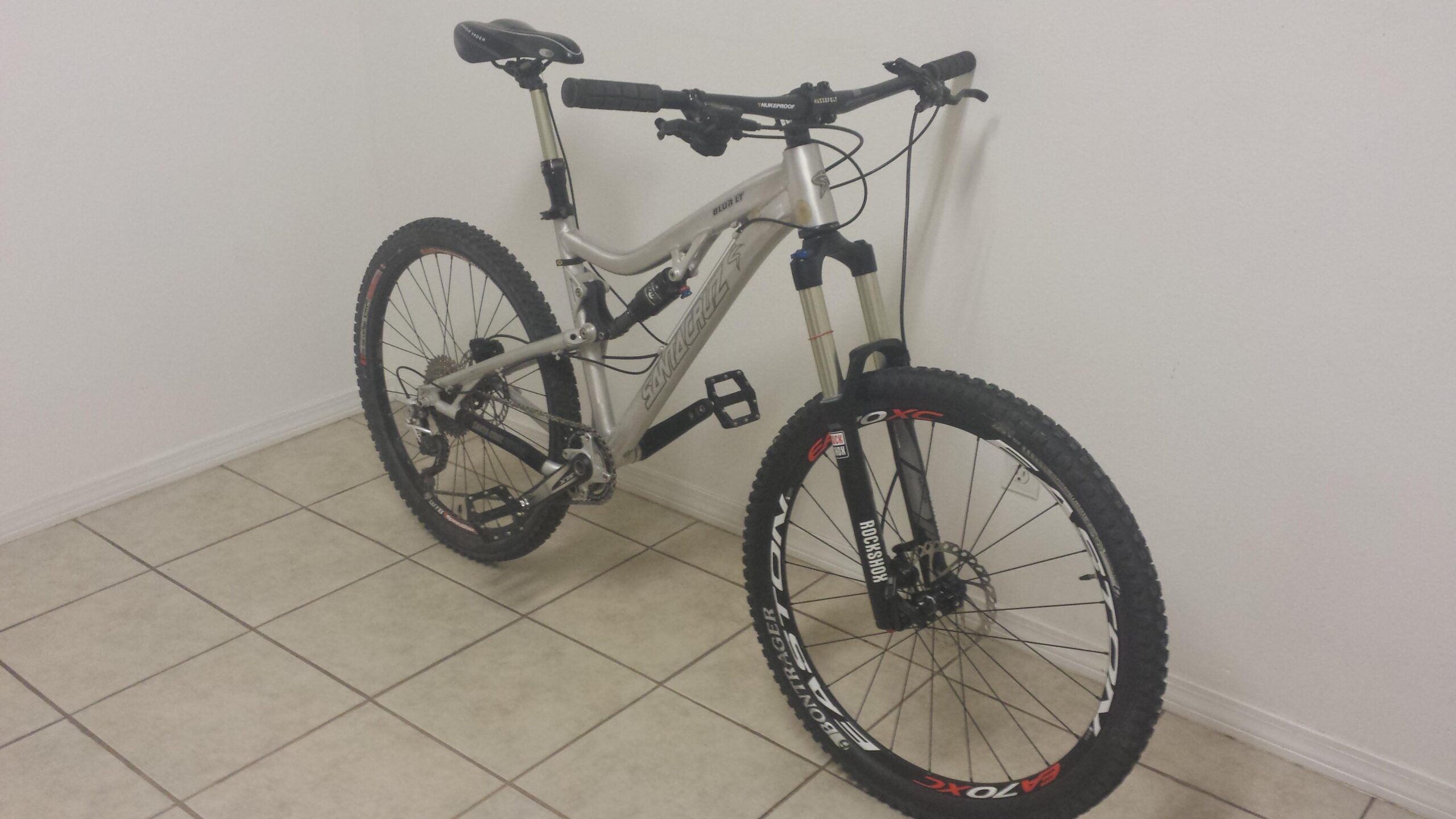 Santa Cruz Blur: A silver mountain bike with front suspension is resting against a white wall. The bike features thick, black tires with visible brand markings, a black saddle, and handlebars with grips. It is positioned on tile flooring.