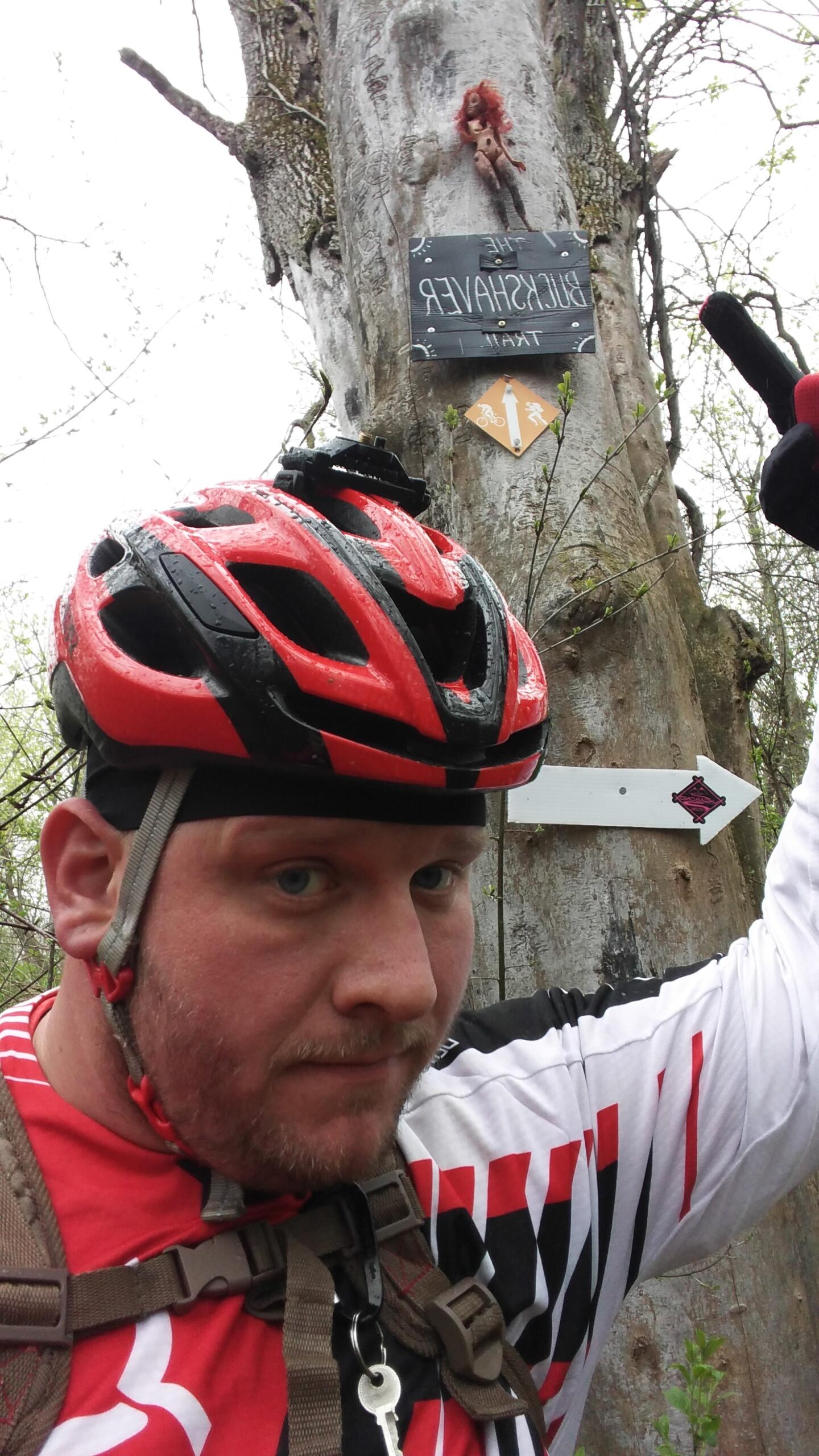A cyclist wearing a red and white helmet and jersey poses in front of a weathered tree. A sign labeled "The Buckshaver Trail" is attached to the tree, along with an arrow pointing in the direction of the trail. A small doll with red hair hangs from the tree above the sign. The setting appears to be a wooded area with overcast skies. Morton-Taylor Trail mountain bike trail.