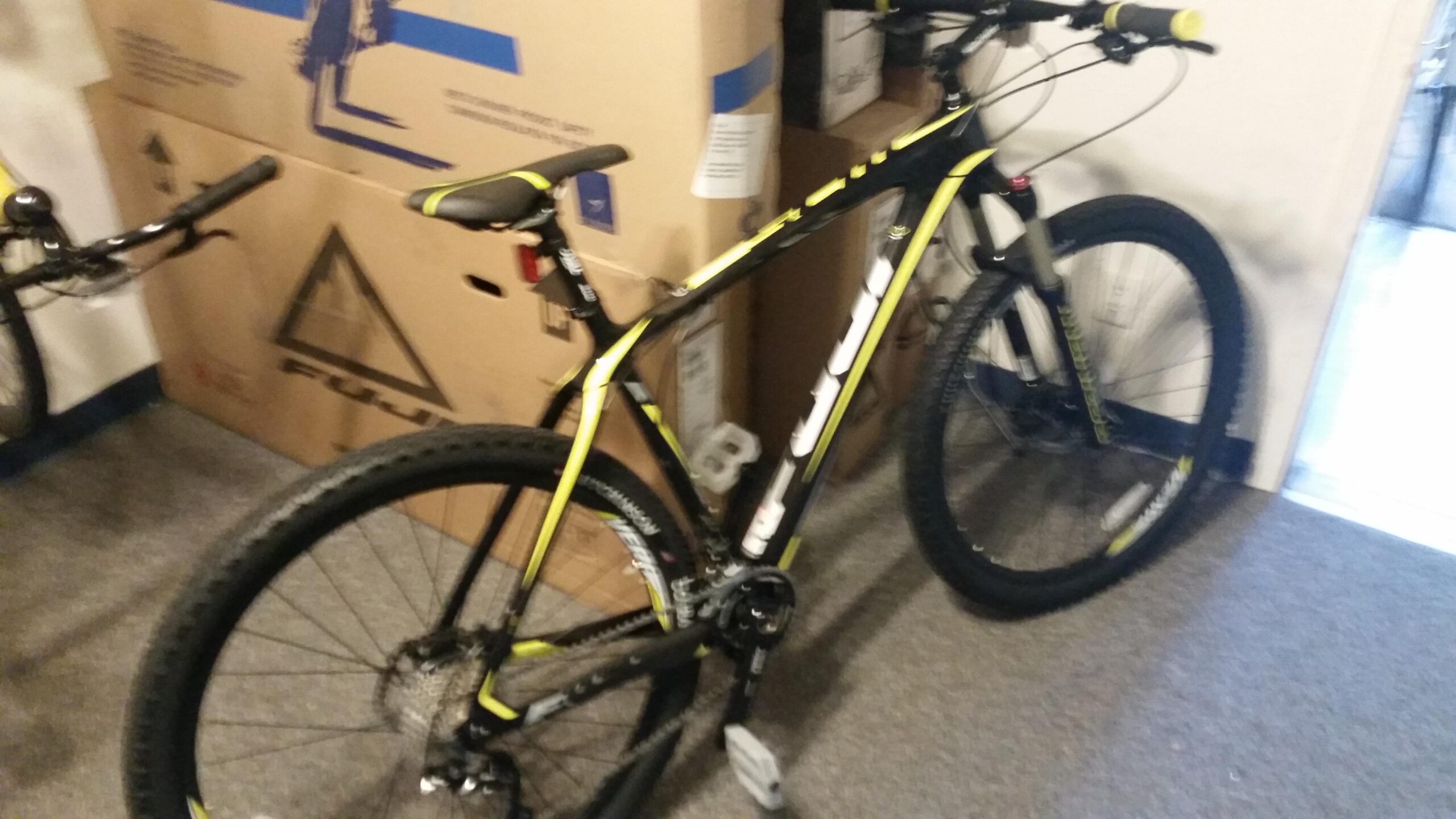 Fuji Fuji SLM 2.5: Alt text: A black and yellow mountain bike positioned in a room with cardboard boxes in the background. The bike features thick tires, a front suspension fork, and a sporty design.