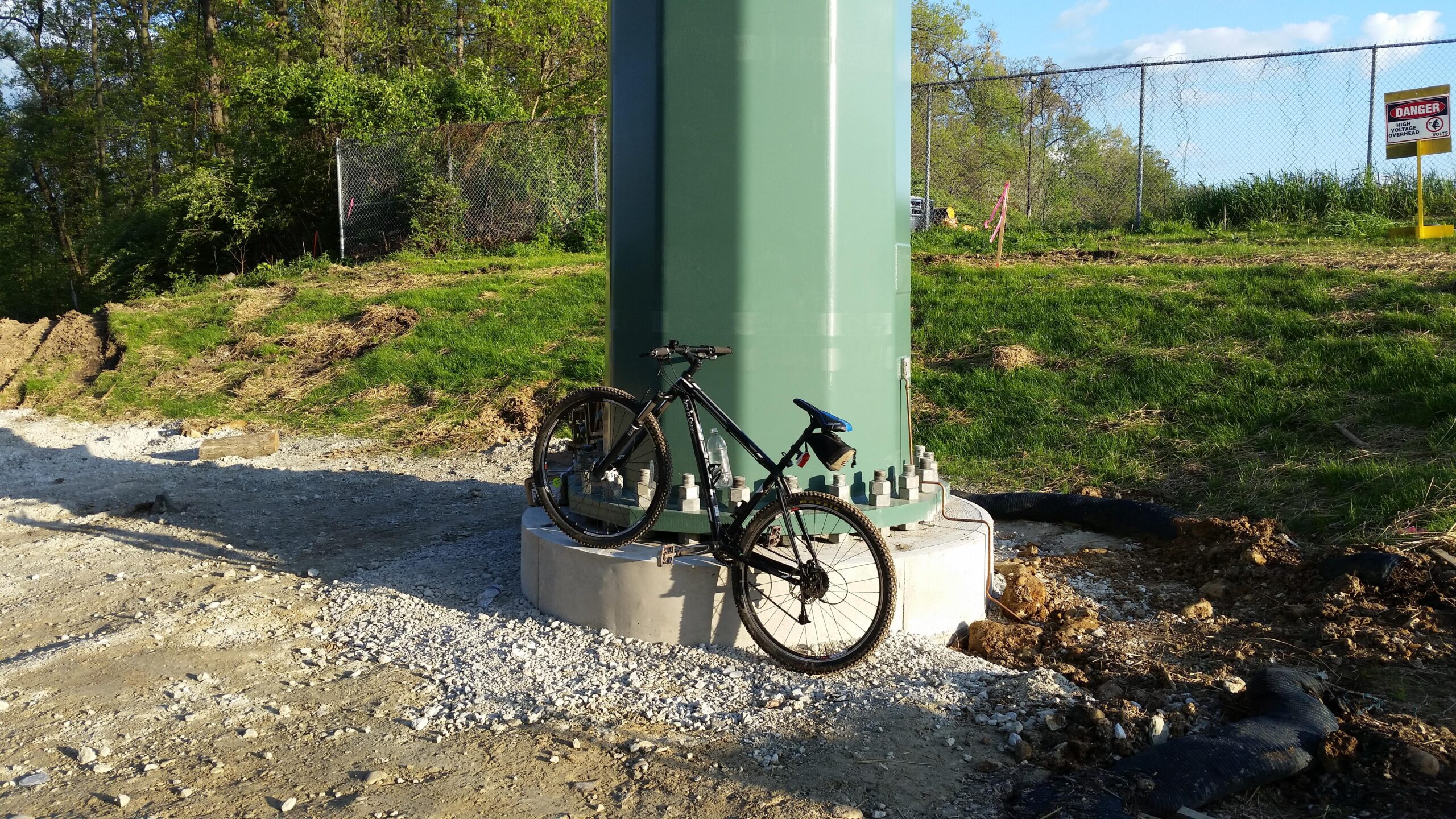 Trek Mamba: A black mountain bike is leaning against a large green cylindrical structure on a gravel path, surrounded by greenery and dirt. In the background, there's a chain-link fence and a warning sign indicating danger.