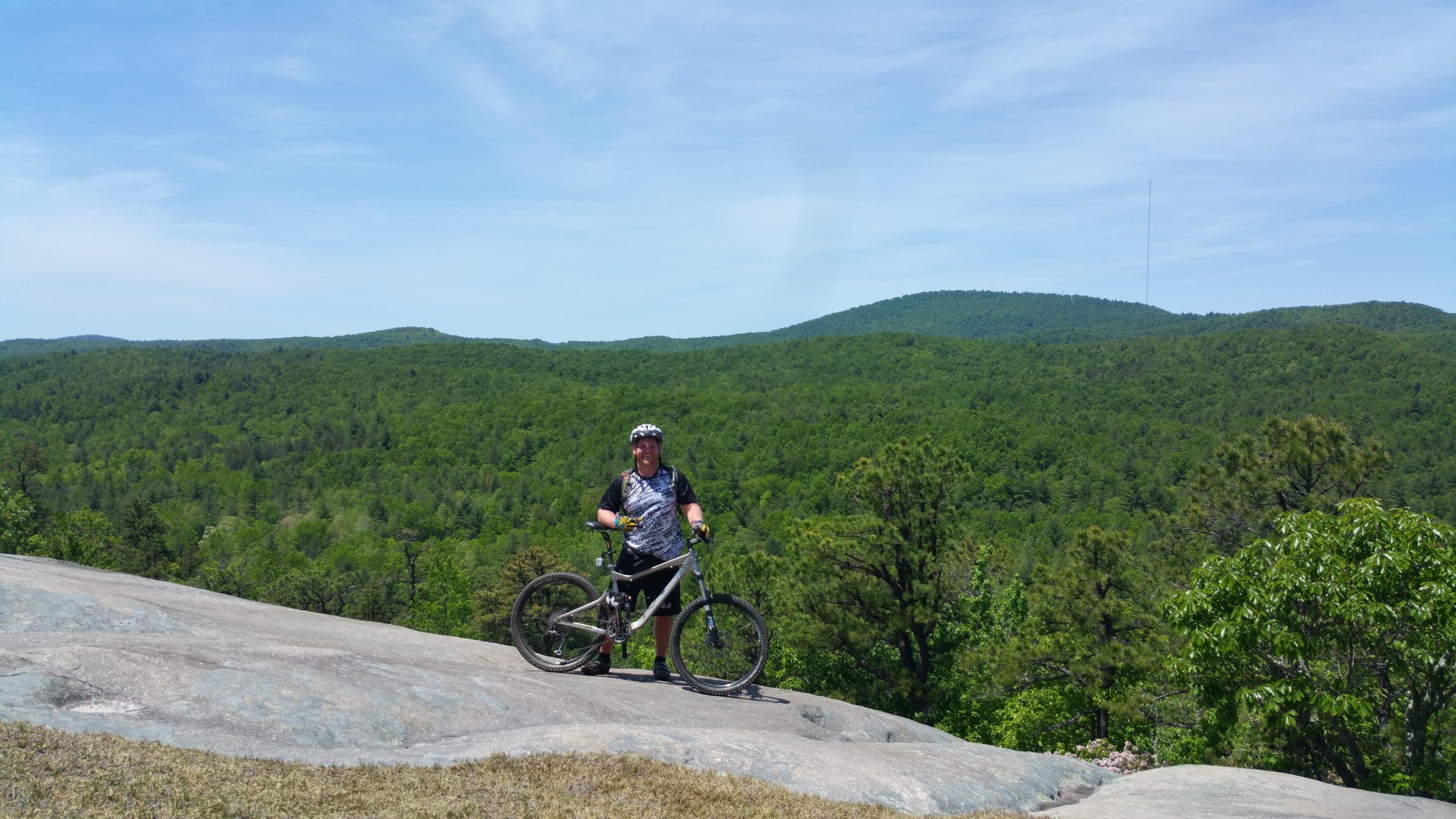 A person standing on a rocky outcrop with a mountain bike, surrounded by lush green hills and trees under a bright blue sky. DuPont State Forest mountain bike trail.