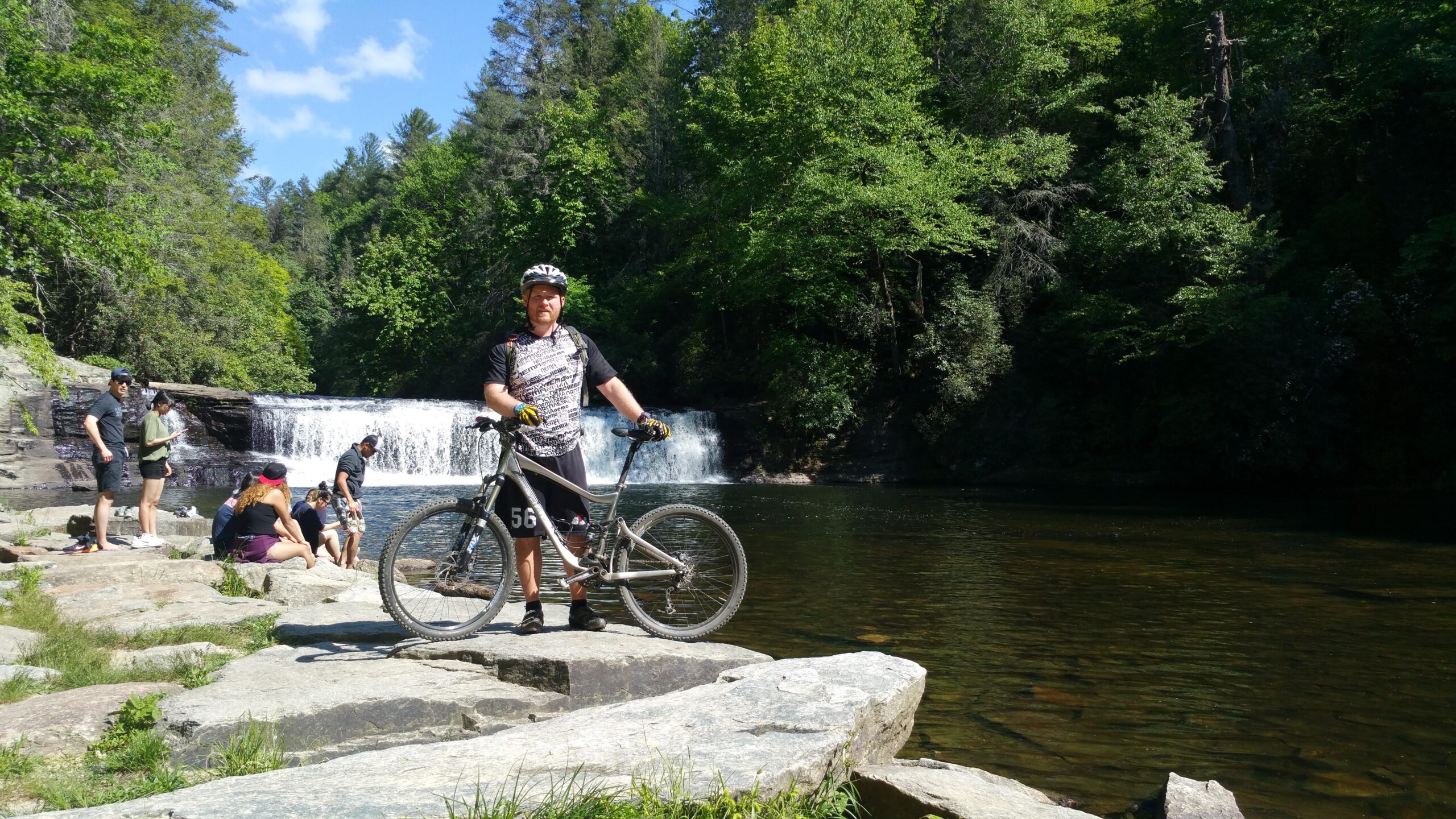 A man stands next to a mountain bike on a rock near a river, with a waterfall and lush green trees in the background. Several people are enjoying the area, some sitting on the rocks and others wading in the water on a sunny day. DuPont State Forest mountain bike trail.