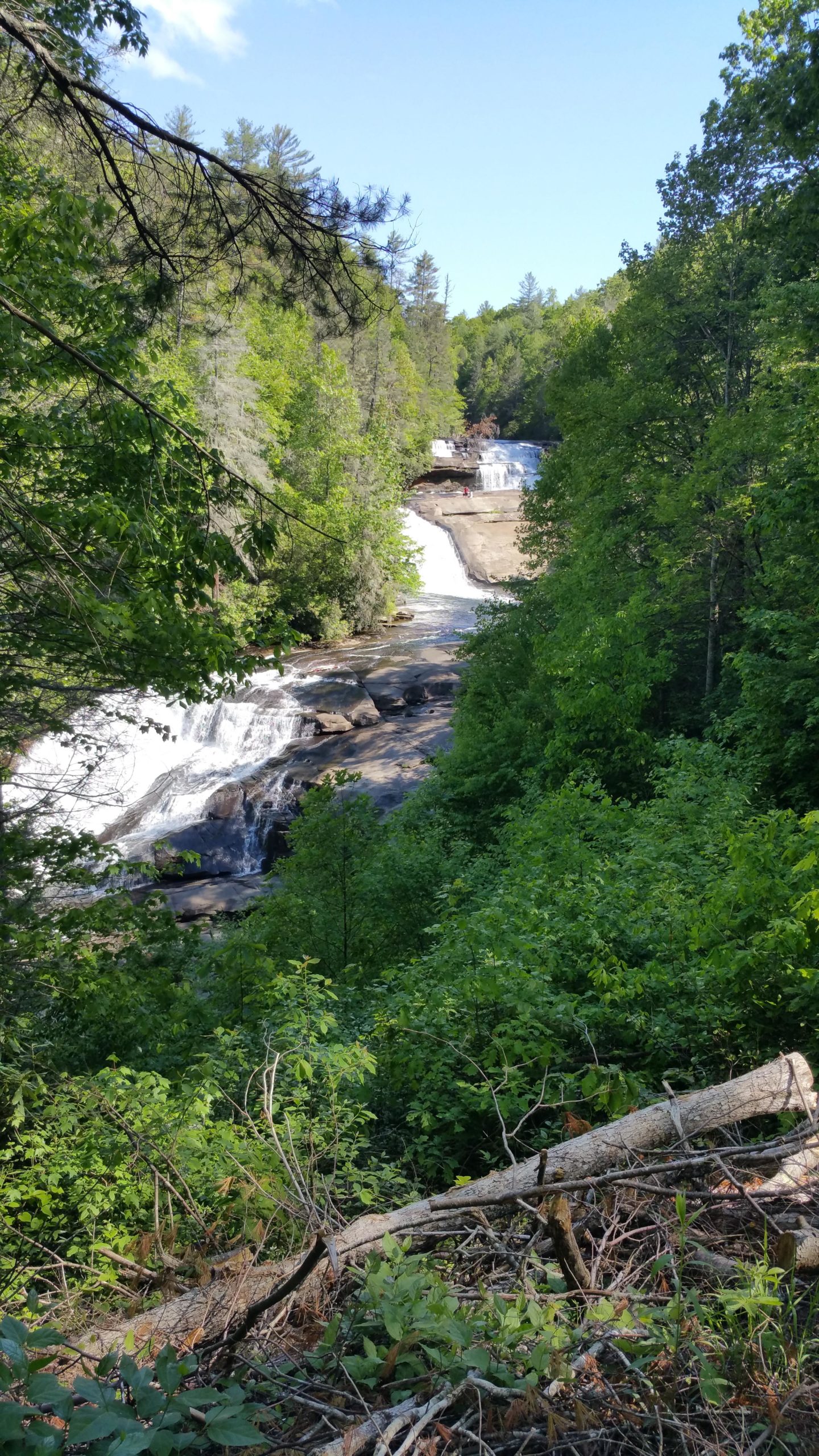 A scenic view of a waterfall cascading over rocks, surrounded by lush greenery and trees. The sky is clear with a few clouds, and a small area of water can be seen flowing below the falls. DuPont State Forest mountain bike trail.