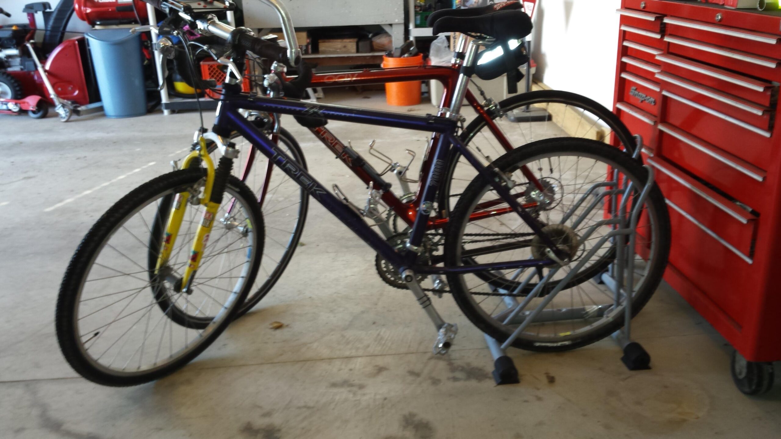 Trek 7000: Two bicycles are parked in a garage, one with yellow front forks and a purple frame, and the other in a red frame. They are positioned next to a bike stand, with a toolbox and various items visible in the background. The garage floor is plain and features some scattered leaves.